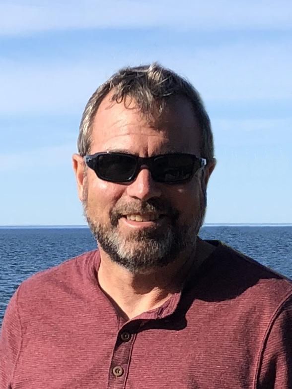 Man with beard and sunglasses smiling in front of a blue sea. He wears a maroon shirt.