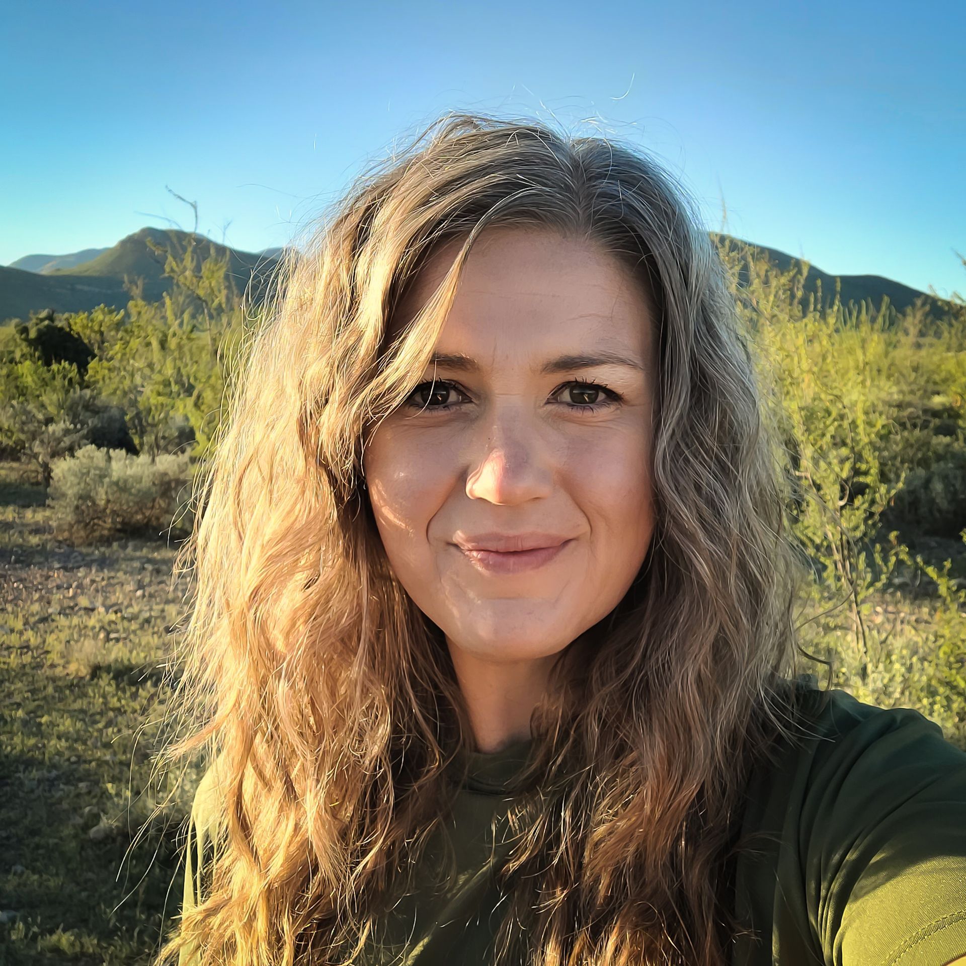 Woman with long curly hair smiles outdoors, backlit by the sun. Mountains and desert vegetation in the background.