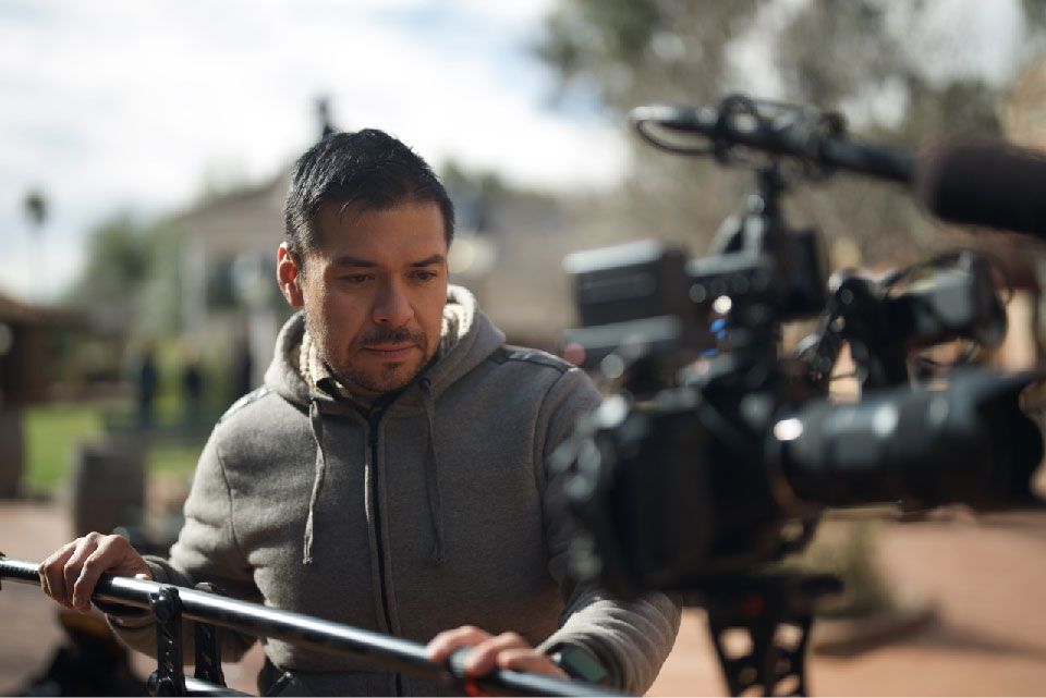 Man operating a film camera on a set, holding a rig. Wearing a gray hoodie, outdoors.