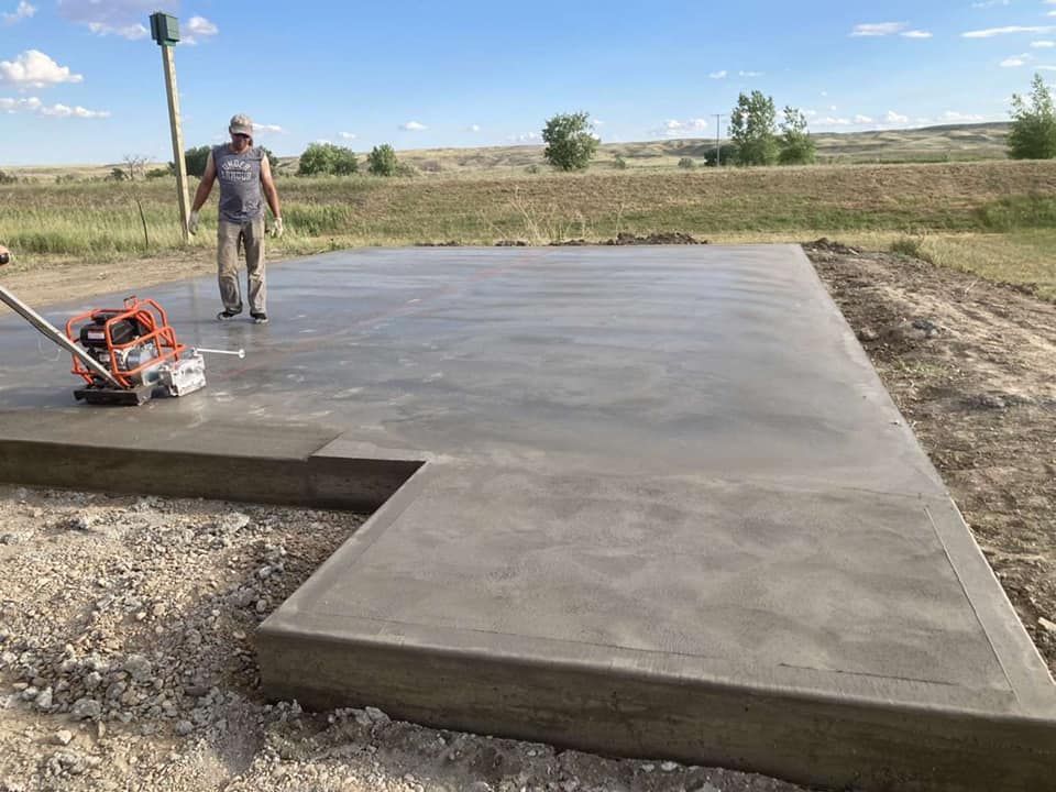 Worker smoothing a freshly poured concrete slab in a rural field with a power trowel.