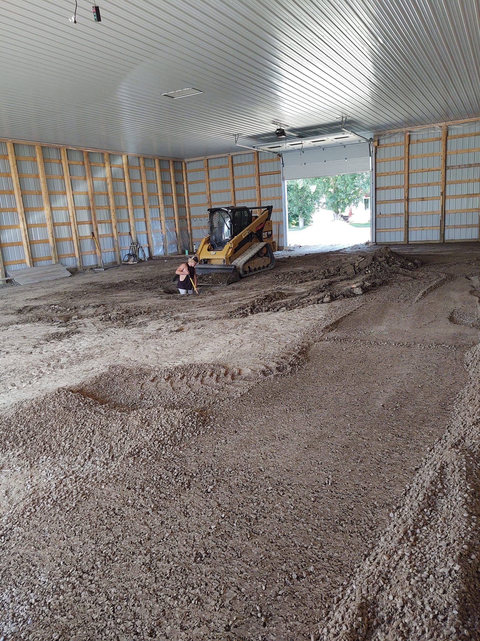 Construction vehicle inside a large unfinished metal building with a gravel floor and open doorway.