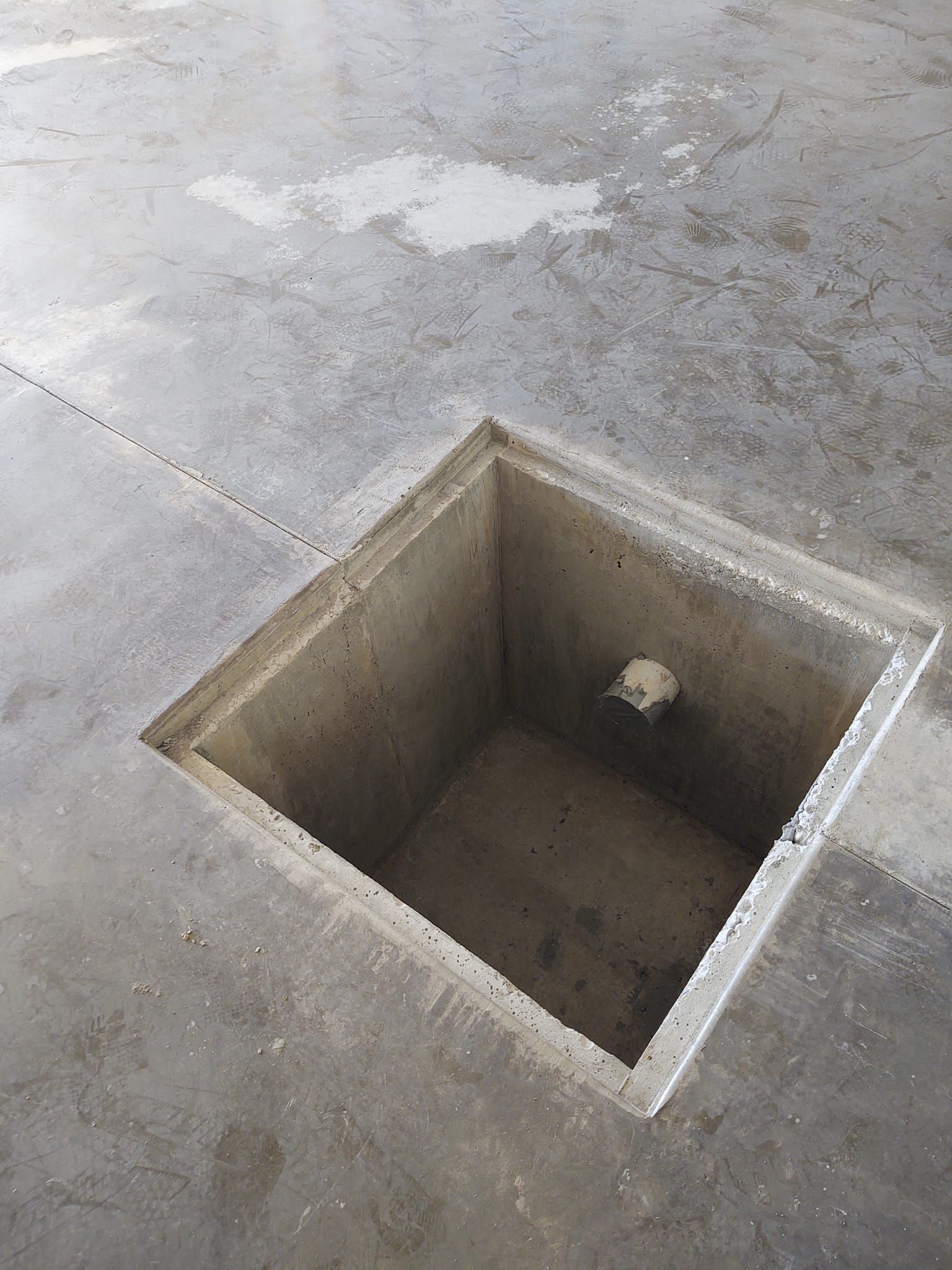 Square concrete floor opening in a gray room, with a small pipe or fixture inside.