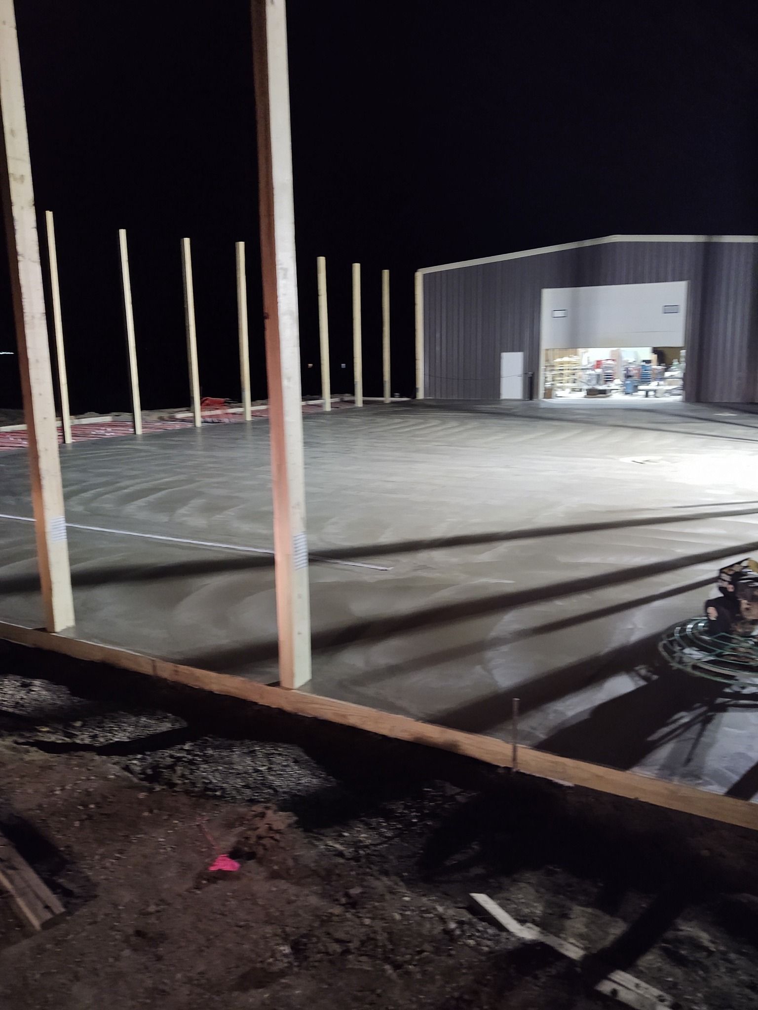 Nighttime construction site with concrete slab, vertical posts, and a lit warehouse opening.