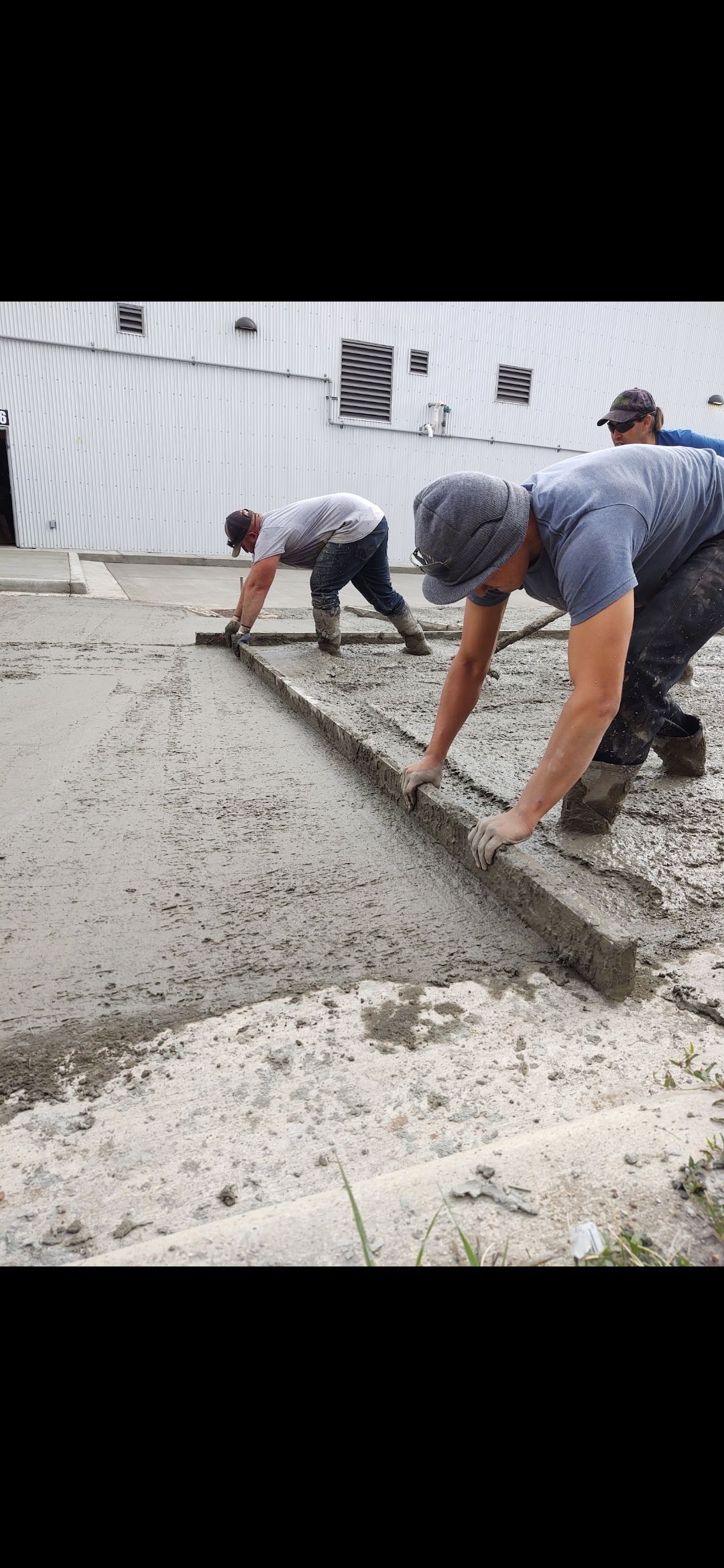 Workers smoothing wet concrete on a driveway outside a building, using long-handled tools.