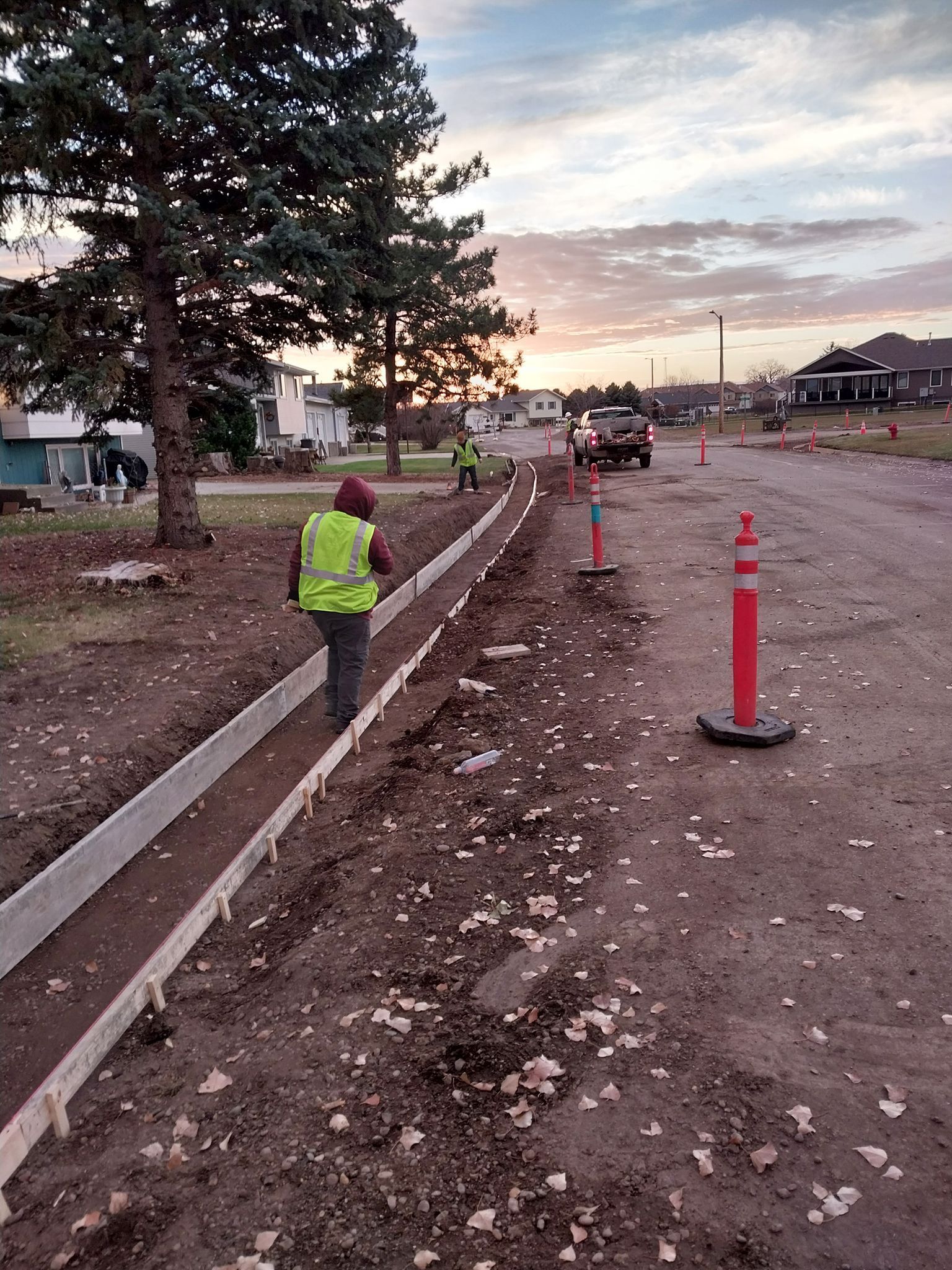 Road construction at dusk with a worker in a yellow vest beside a newly poured curb and traffic cones.