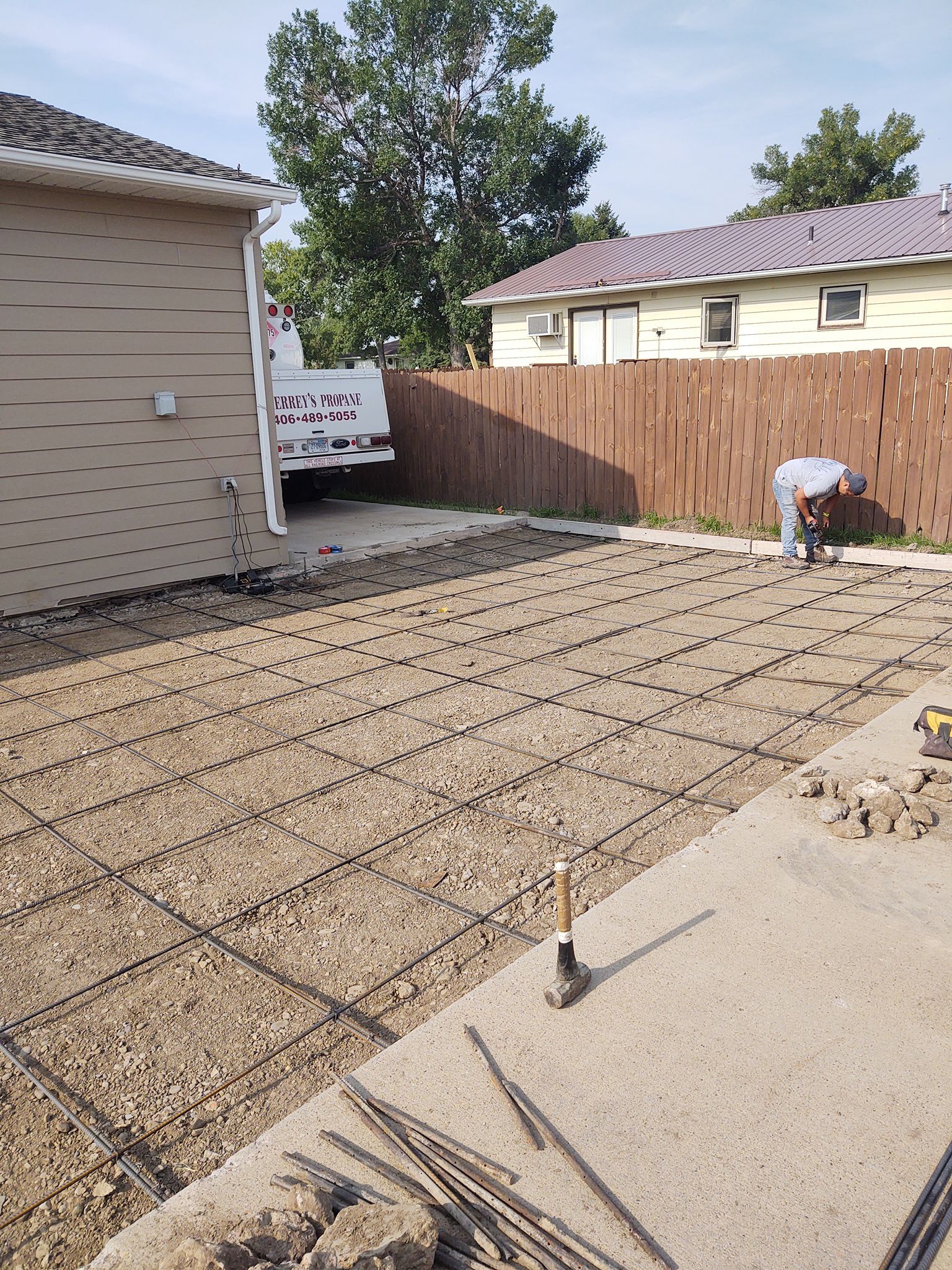 Backyard construction site with rebar grid, concrete slab edges, and a worker near a wooden fence.