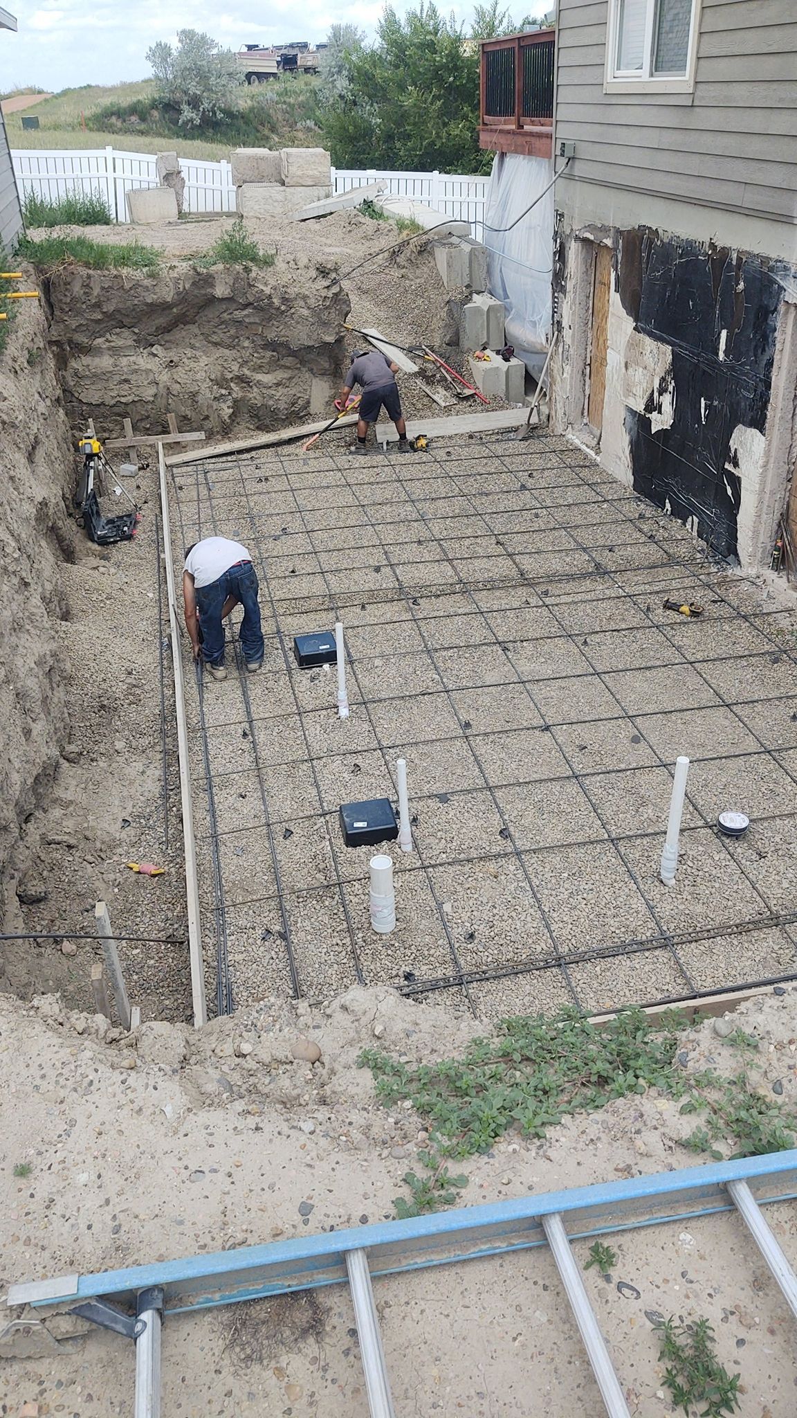 Workers preparing a concrete foundation in a narrow backyard beside a house, with rebar and forms laid out.