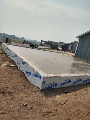 Concrete slab foundation under construction on a bare lot, with protective blue-and-white wrapping along the edge.