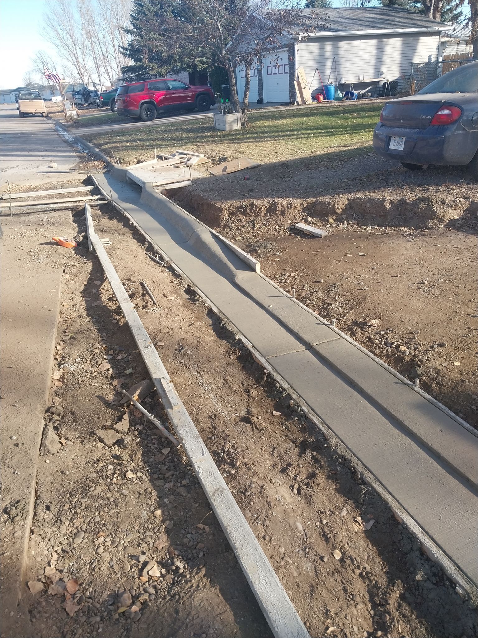 Damaged curb ramp and broken sidewalk beside a residential street with parked cars and houses.