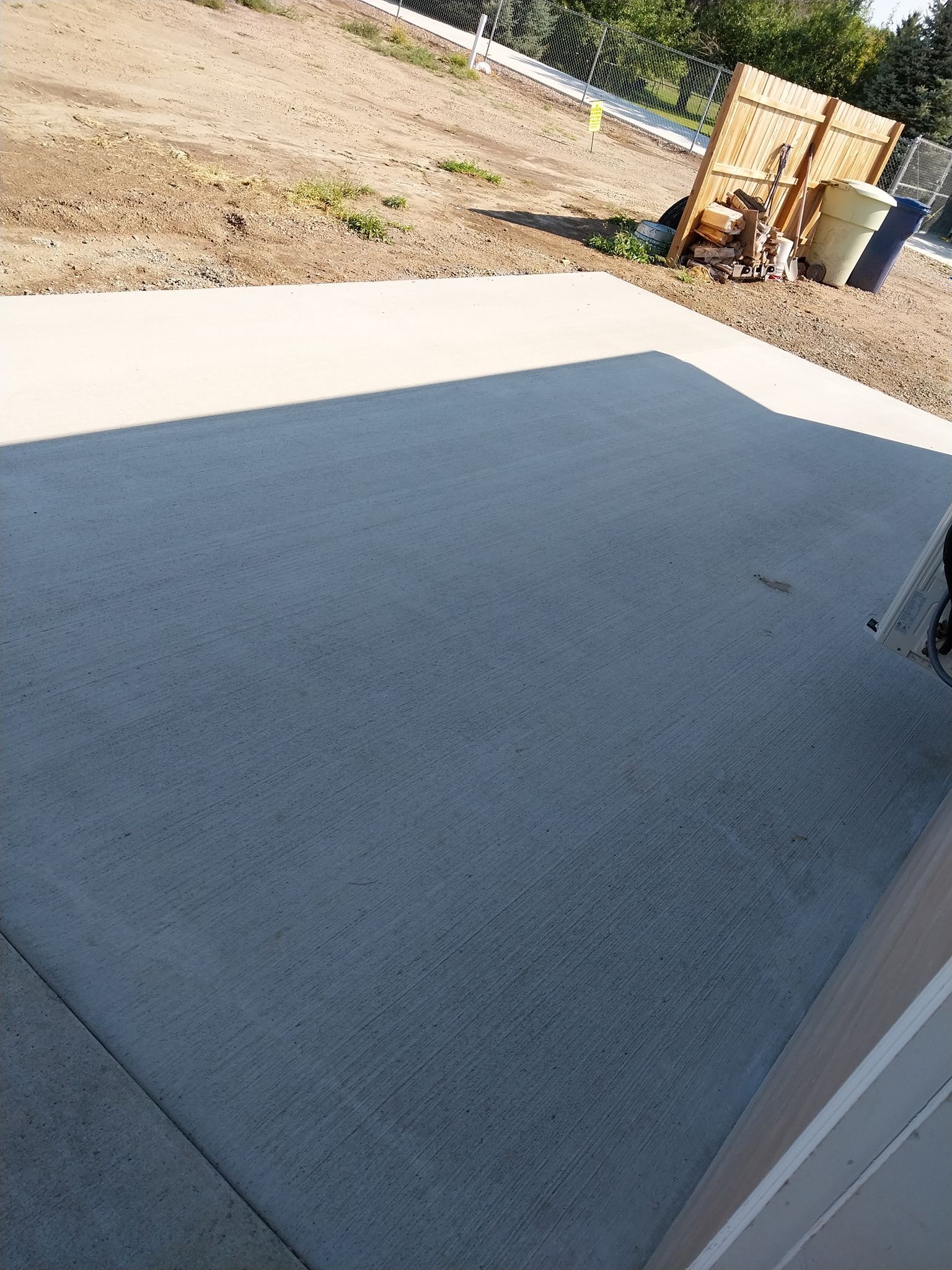 Concrete patio with dark gray coating beside a dirt yard and stacked items in the background.