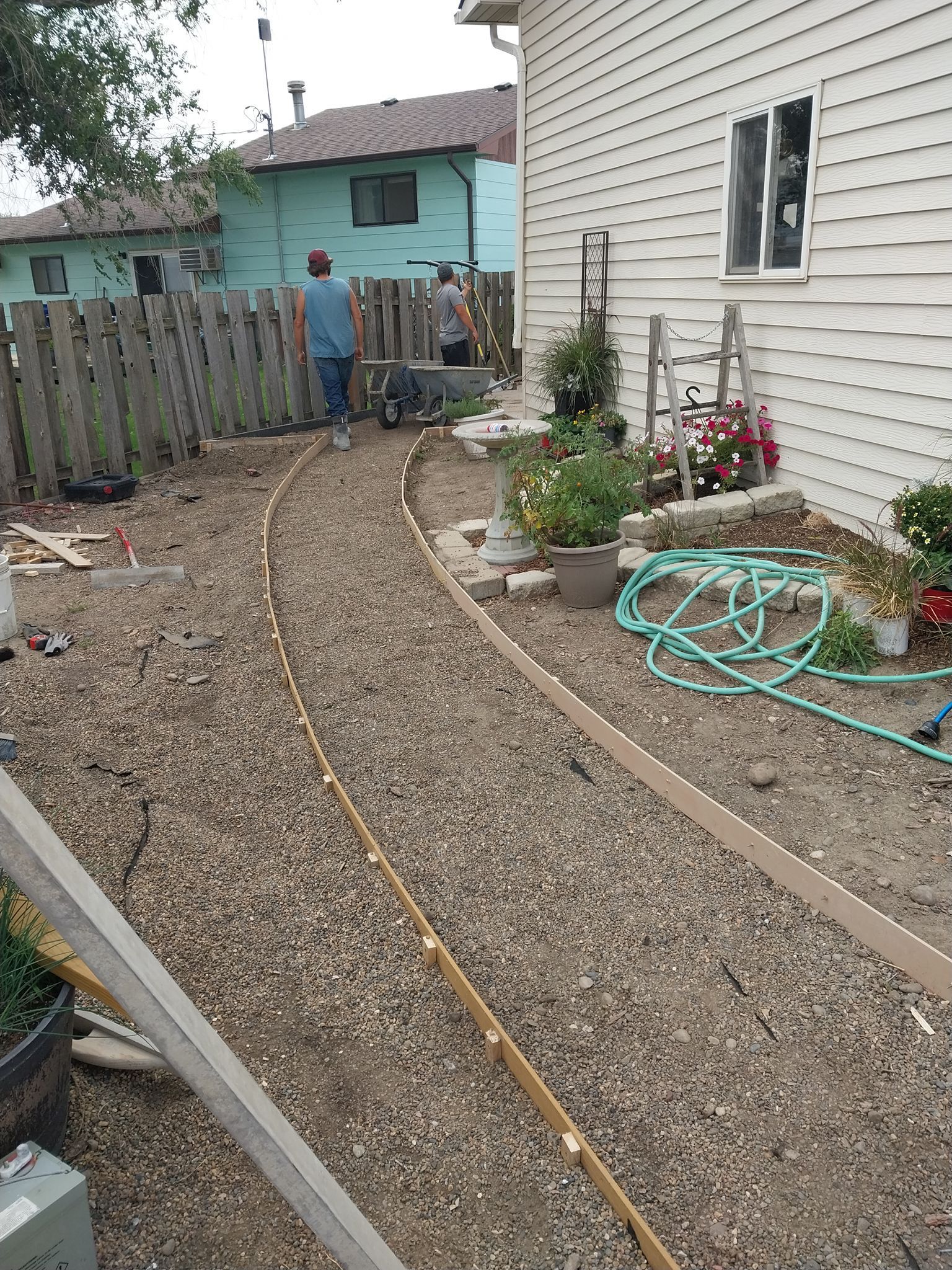 Gravel backyard beside a white house, with a turquoise shed, garden beds, and a coiled green hose.