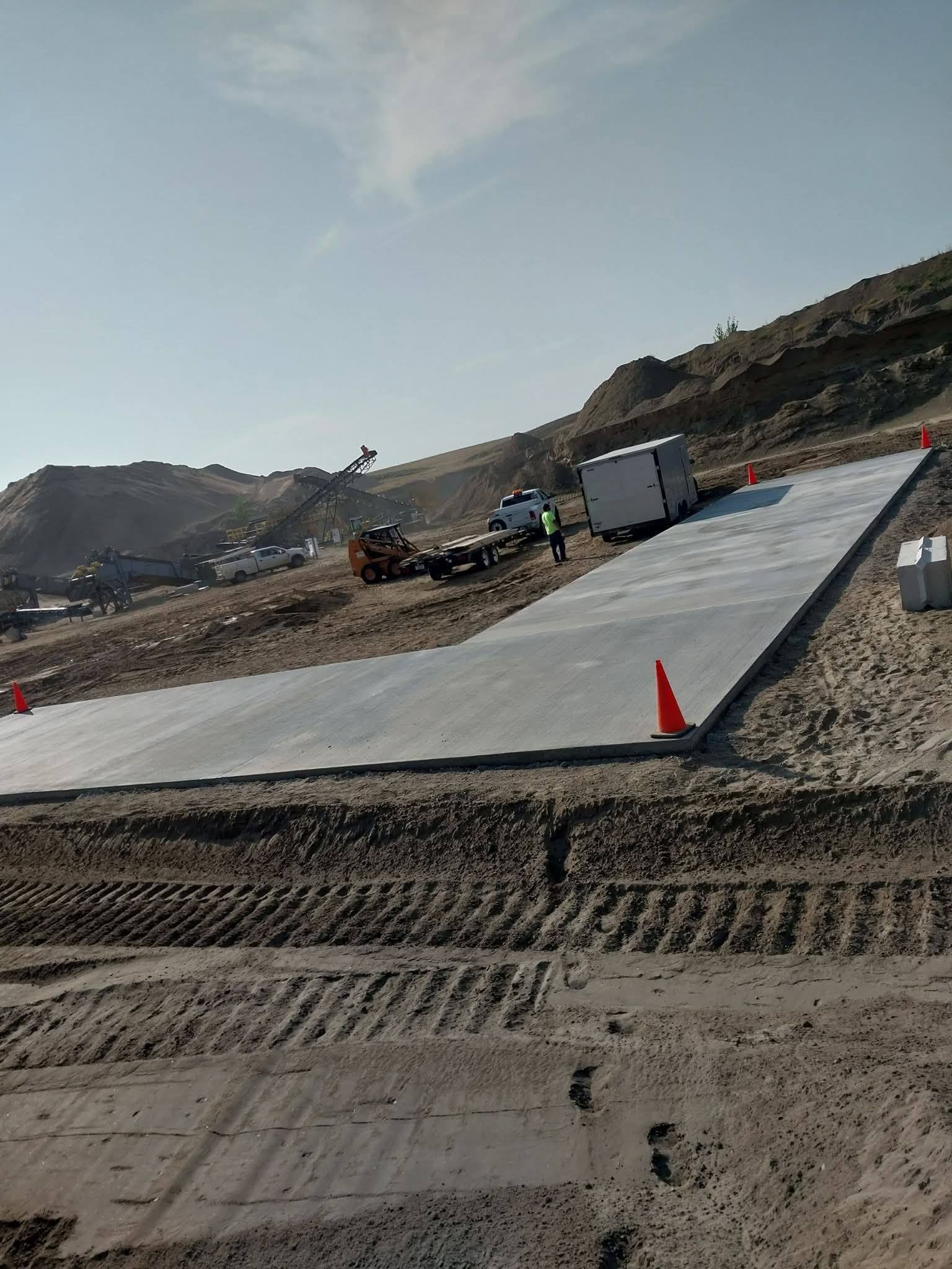 Concrete ramp under construction with orange cones, workers, and equipment on a rocky site under a clear sky