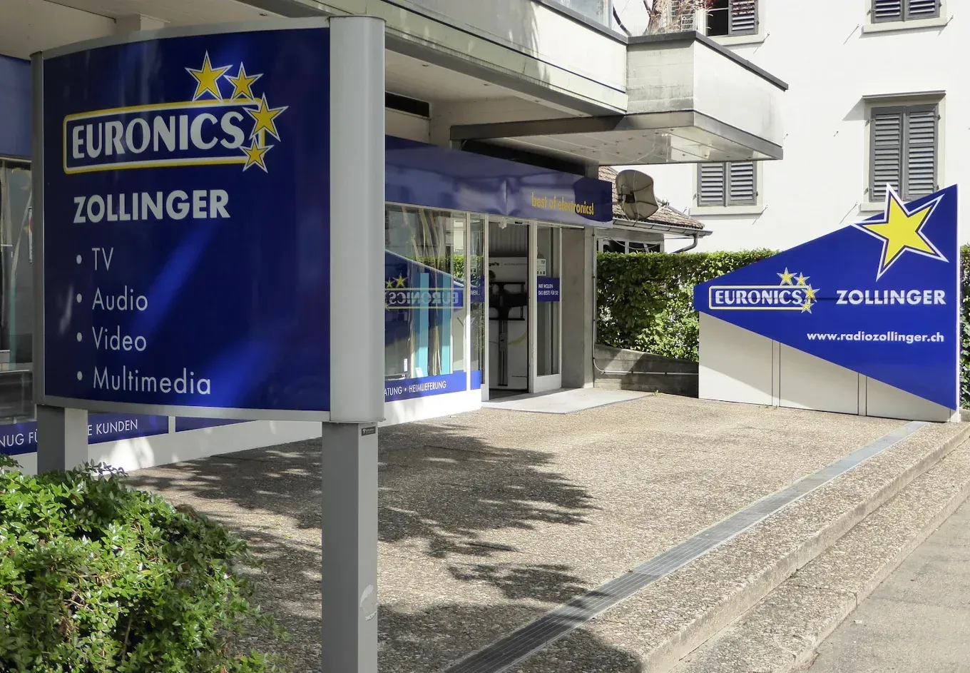 Euronics Zollinger-Geschäftsfront mit blauen Schildern und gläsernem Eingang an einem Kopfsteinpflasterweg
