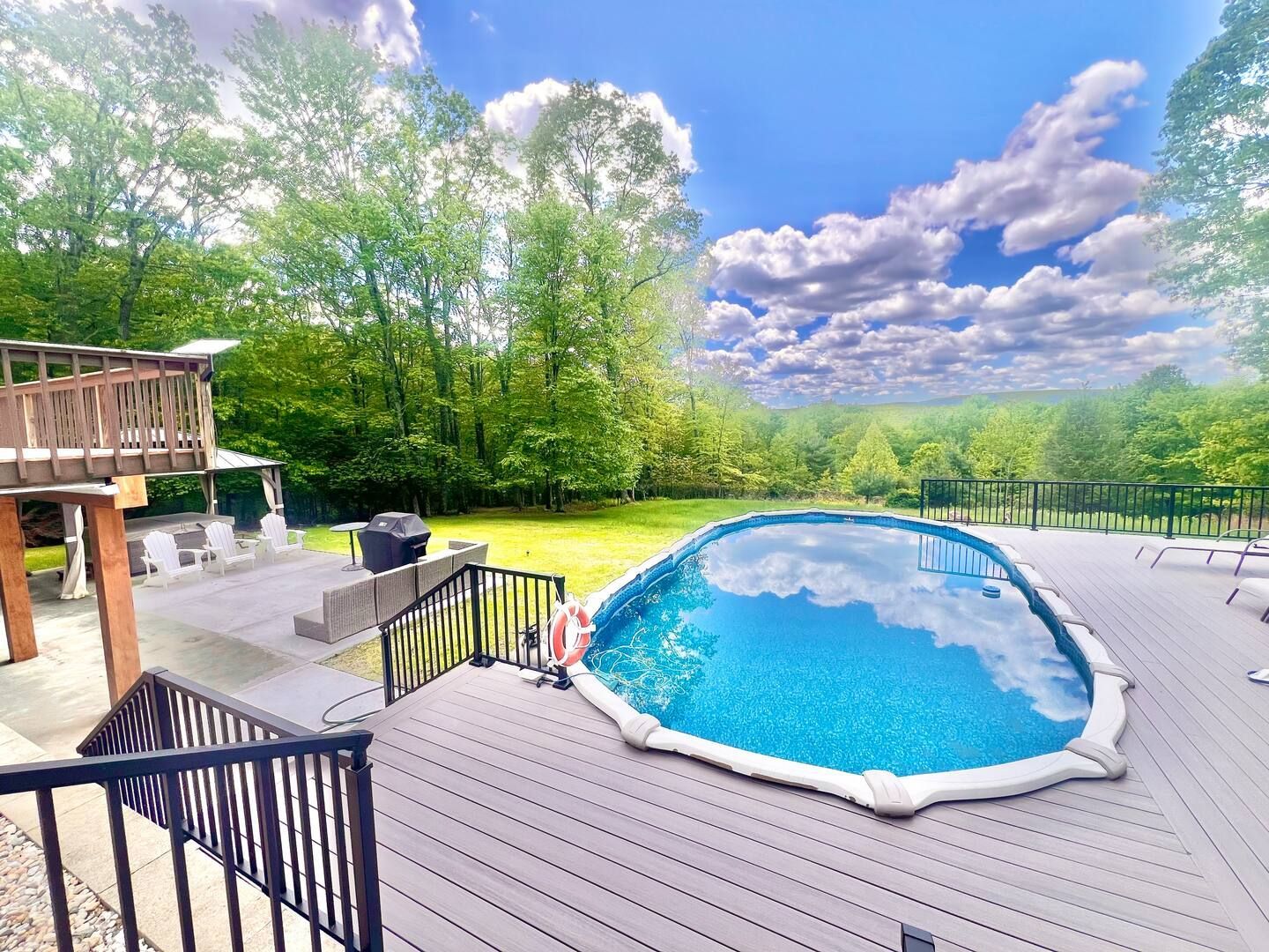 Pool area with blue water, surrounded by a wooden deck, green lawn, trees, and a bright cloudy sky.