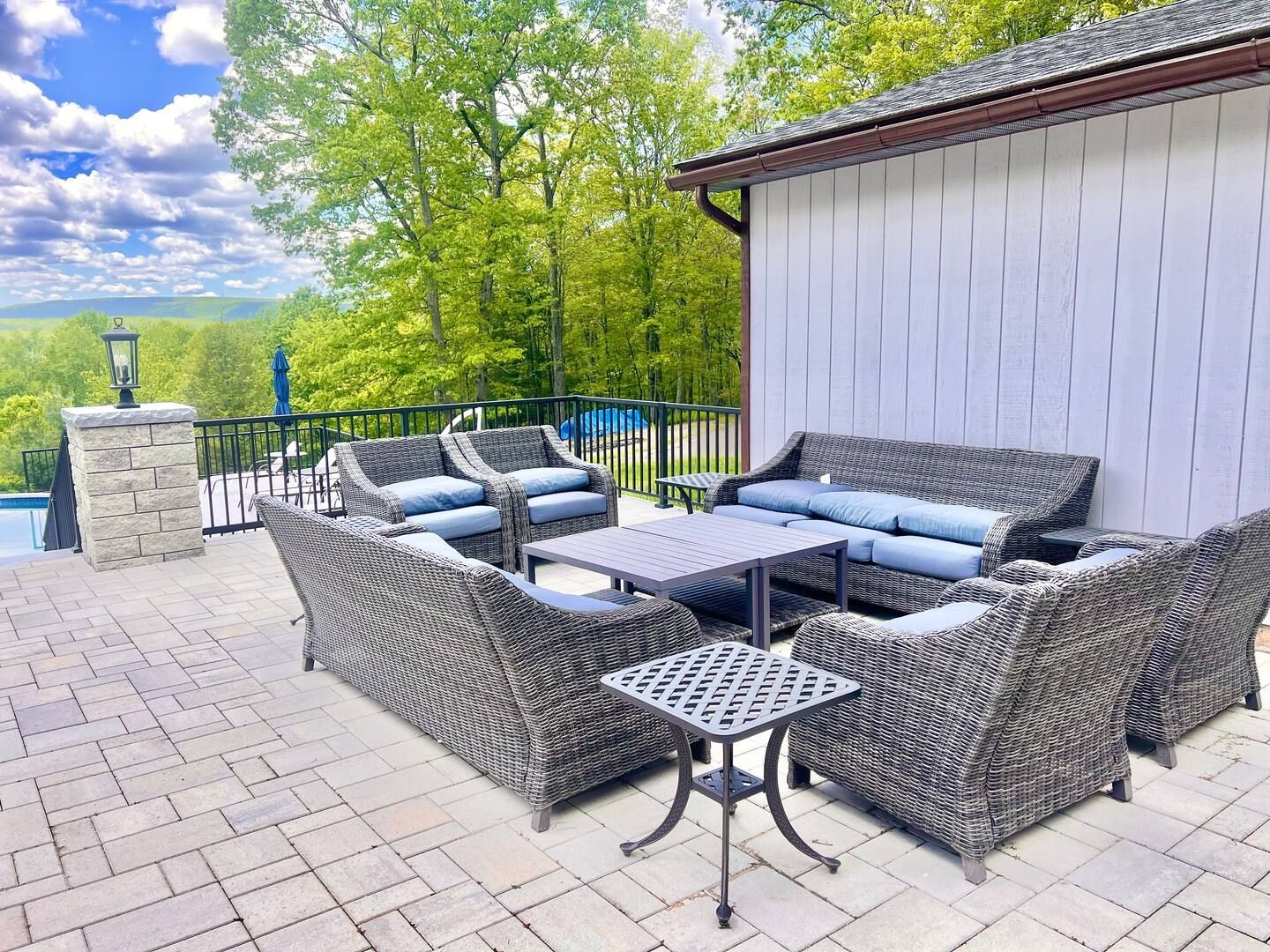 Patio with gray wicker furniture, blue cushions, and a small table. Green trees and blue sky in the background.