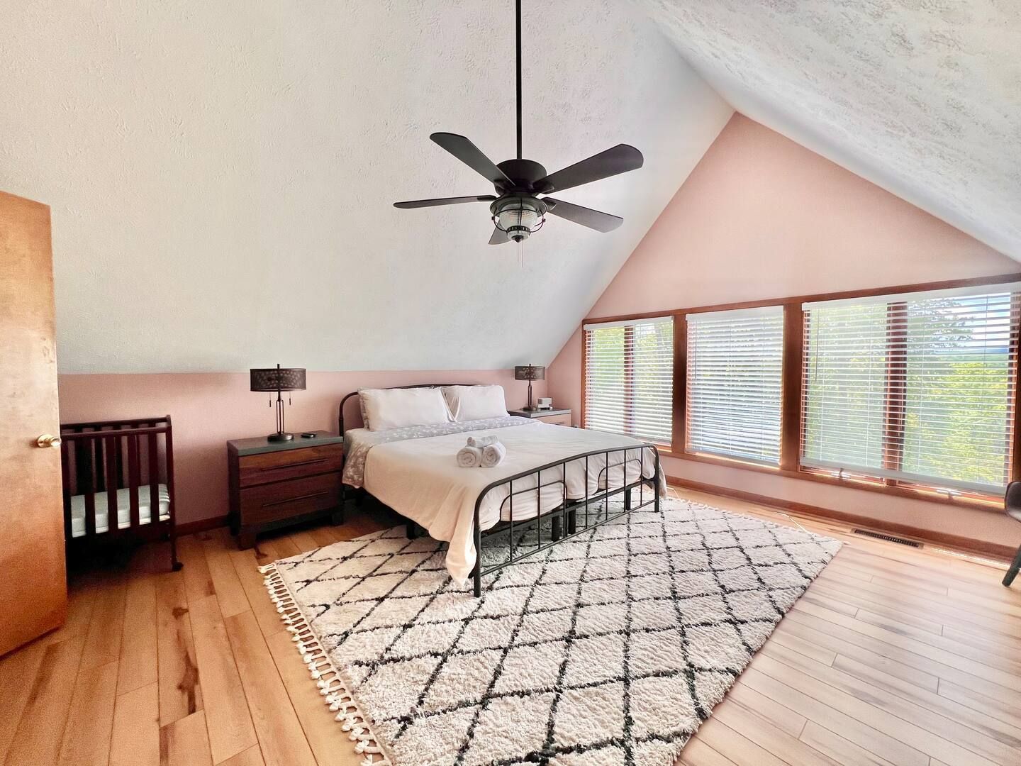 Bedroom with pink walls, hardwood floors, and a large rug. There is a bed, nightstands, and a crib.