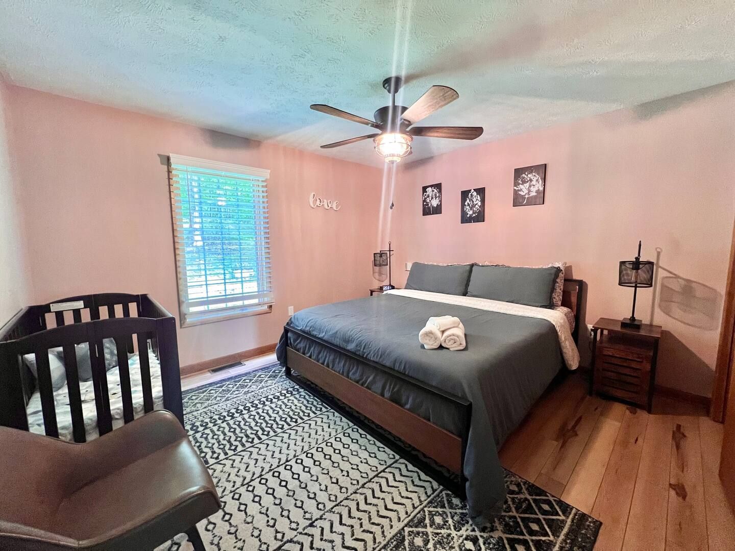Bedroom with a queen-sized bed, crib, and patterned rug on hardwood flooring. Pink walls with framed photos.