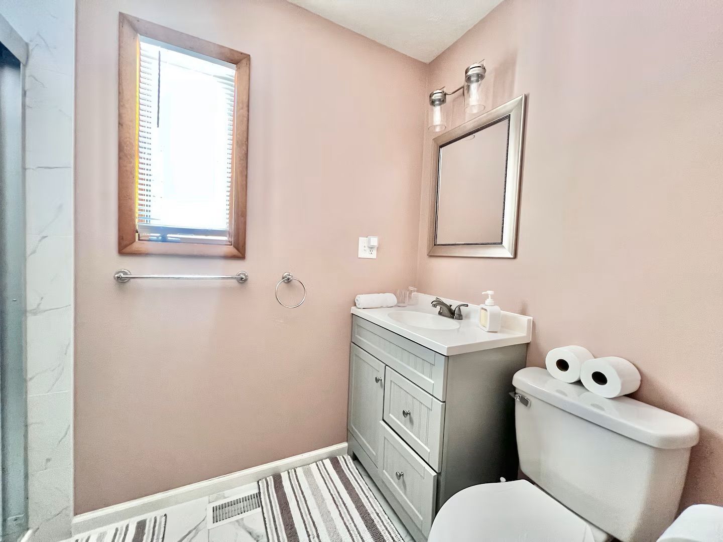Small bathroom with pink walls, gray vanity, and toilet. Window with blinds; striped floor mat.