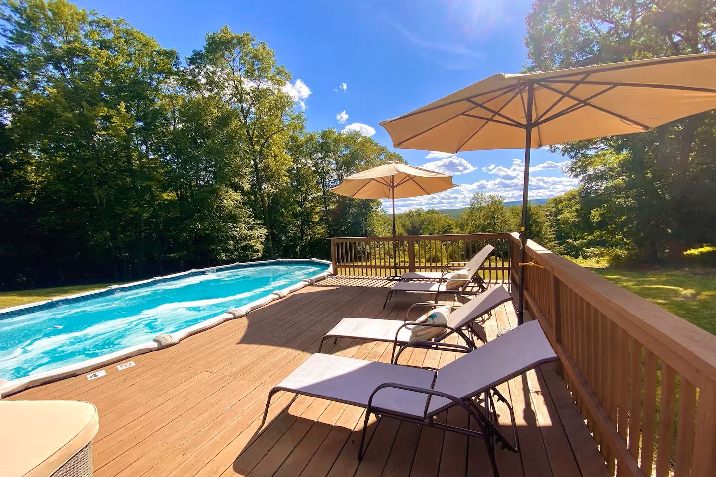 Wooden deck with pool, lounge chairs, and umbrellas in sunny backyard setting.