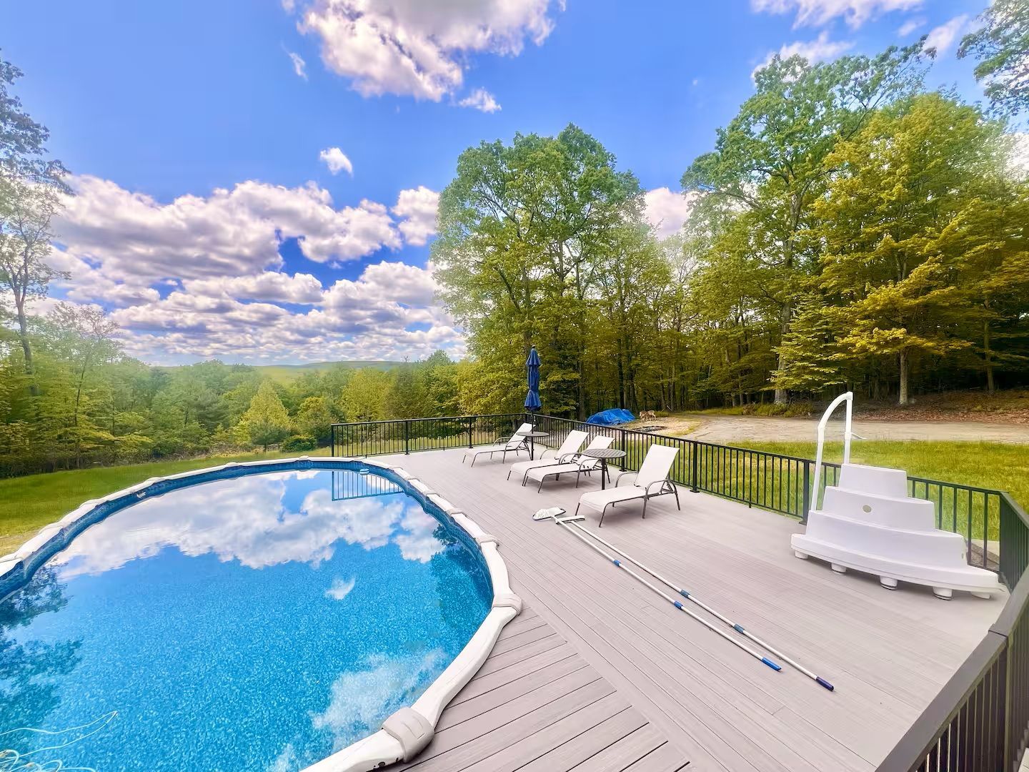 Swimming pool with deck, lounge chairs, and wooded backdrop under a blue sky with clouds.