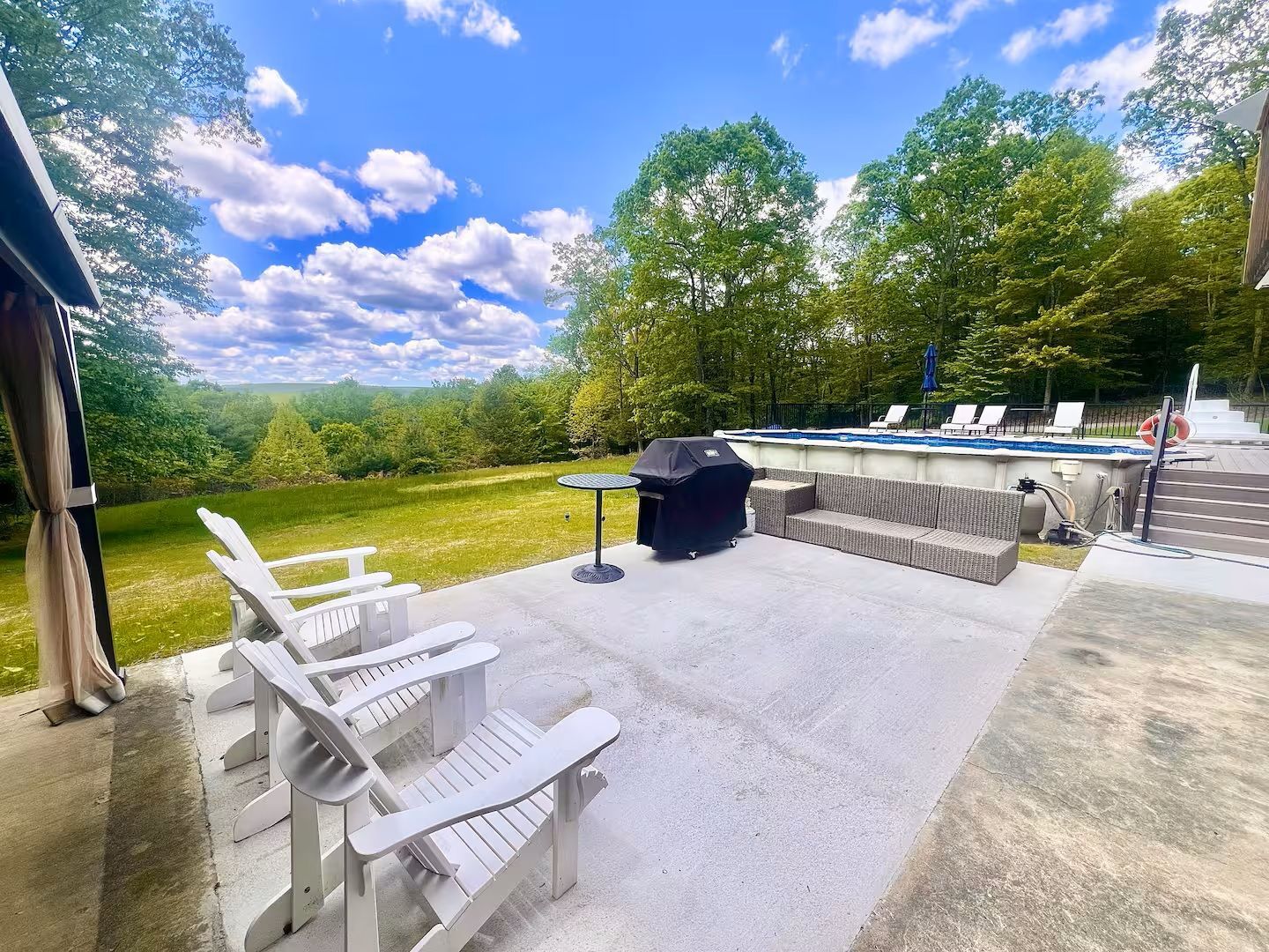 Patio with white chairs, grill, and pool overlooking a green landscape under a blue sky.