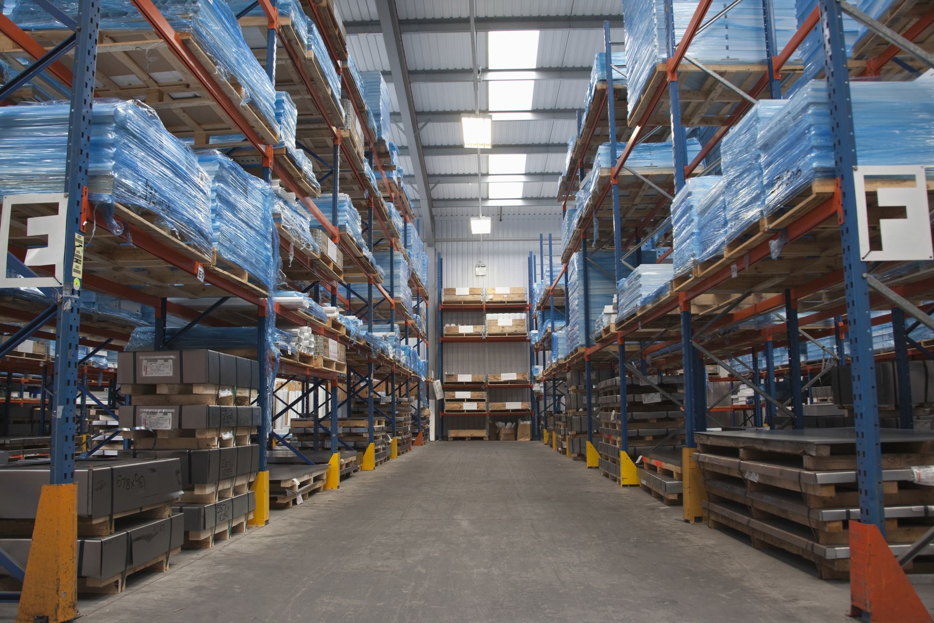 Warehouse aisle with tall metal shelving stacked with blue-wrapped pallets and boxes.