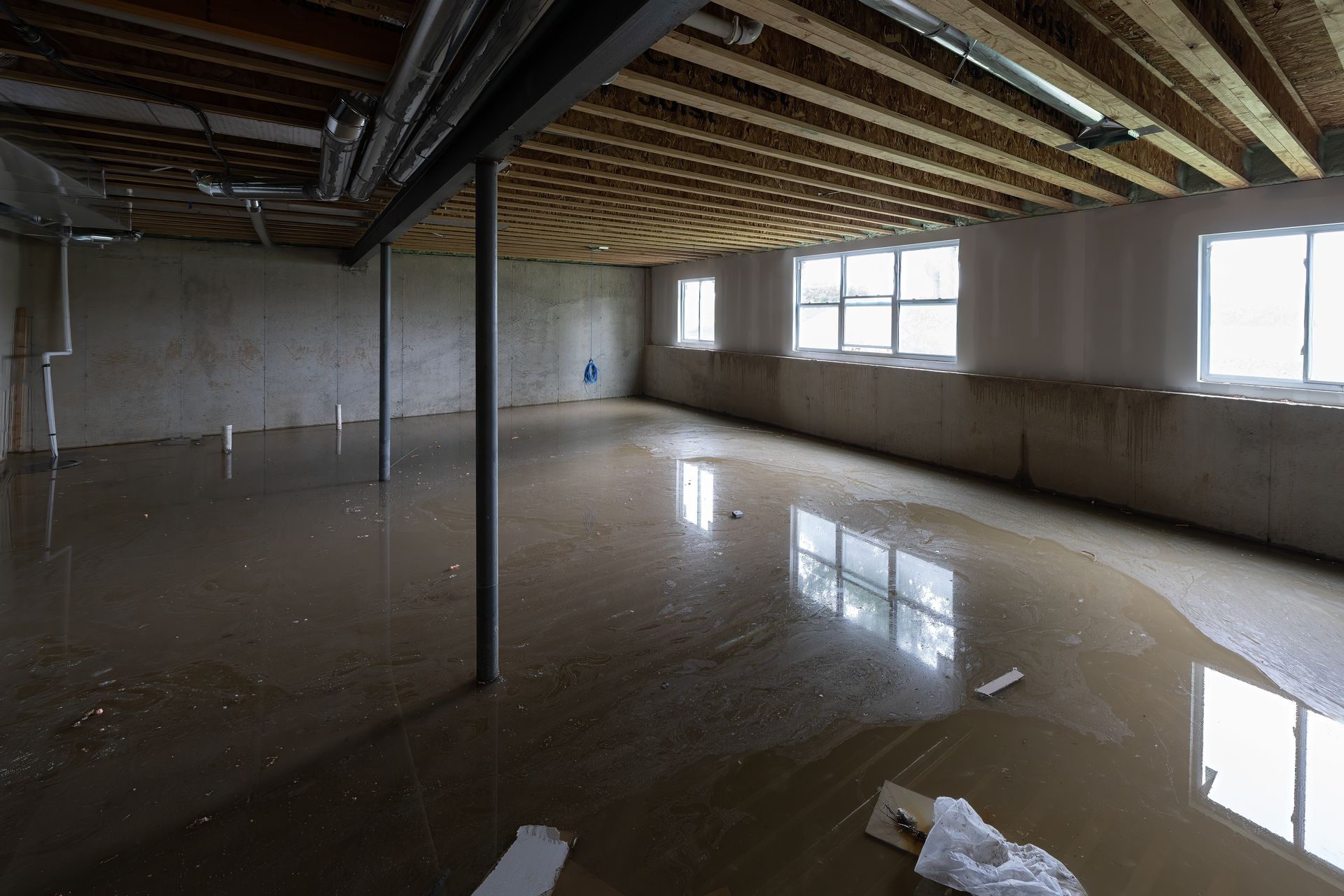 Flooded unfinished basement with exposed beams and windows, reflecting water covering the floor