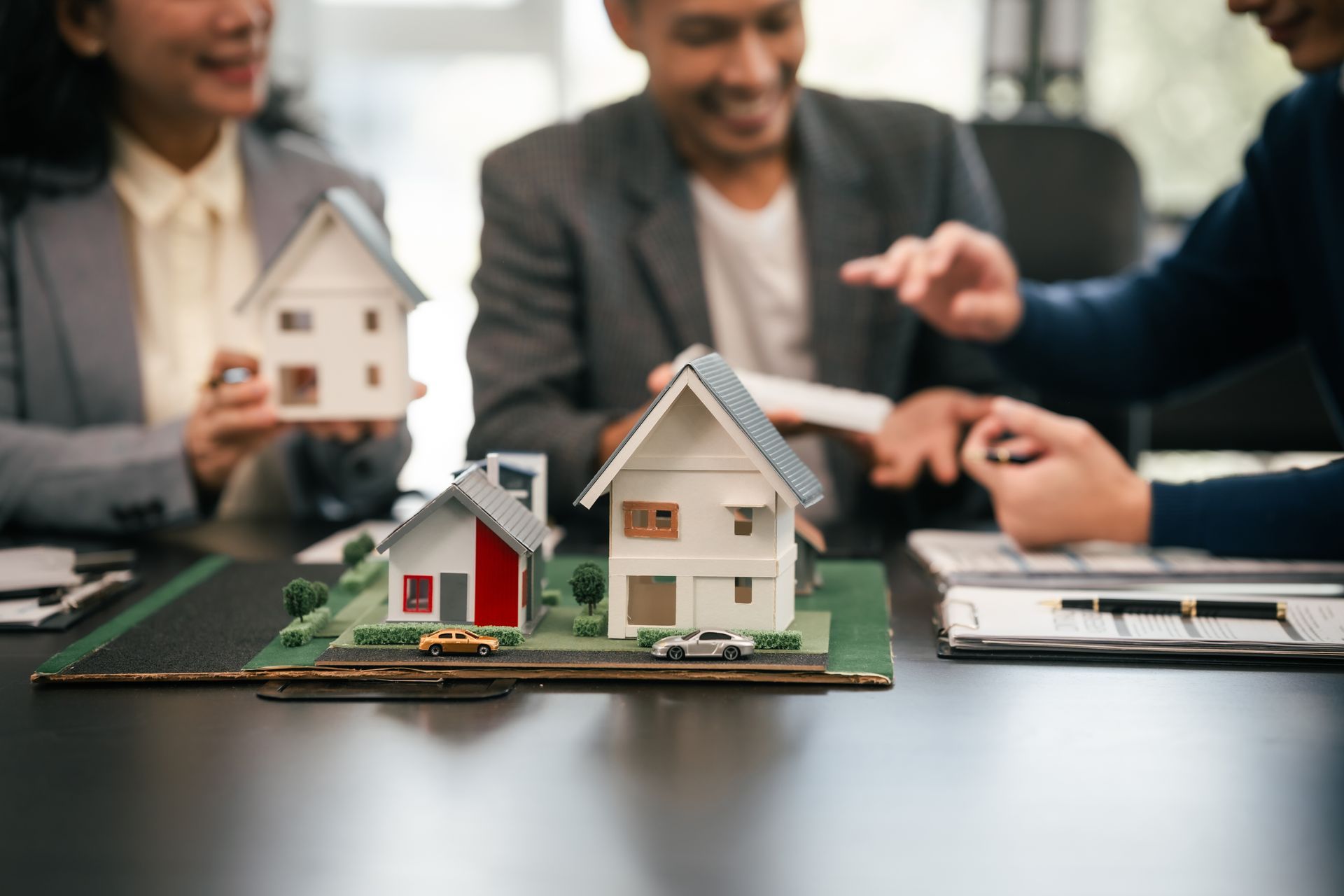 Miniature house models on a table during a real estate meeting, with people discussing in the background