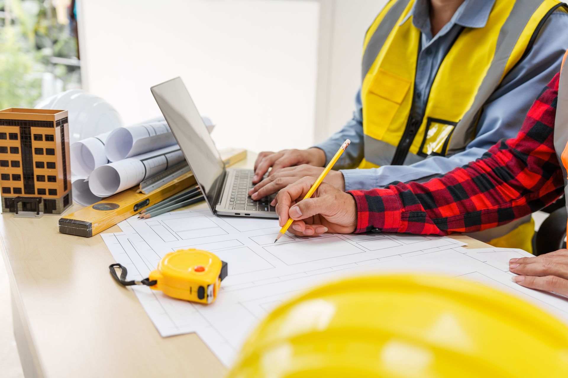Construction workers reviewing plans at a desk with laptop, hard hats, measuring tape, and rolled blueprints