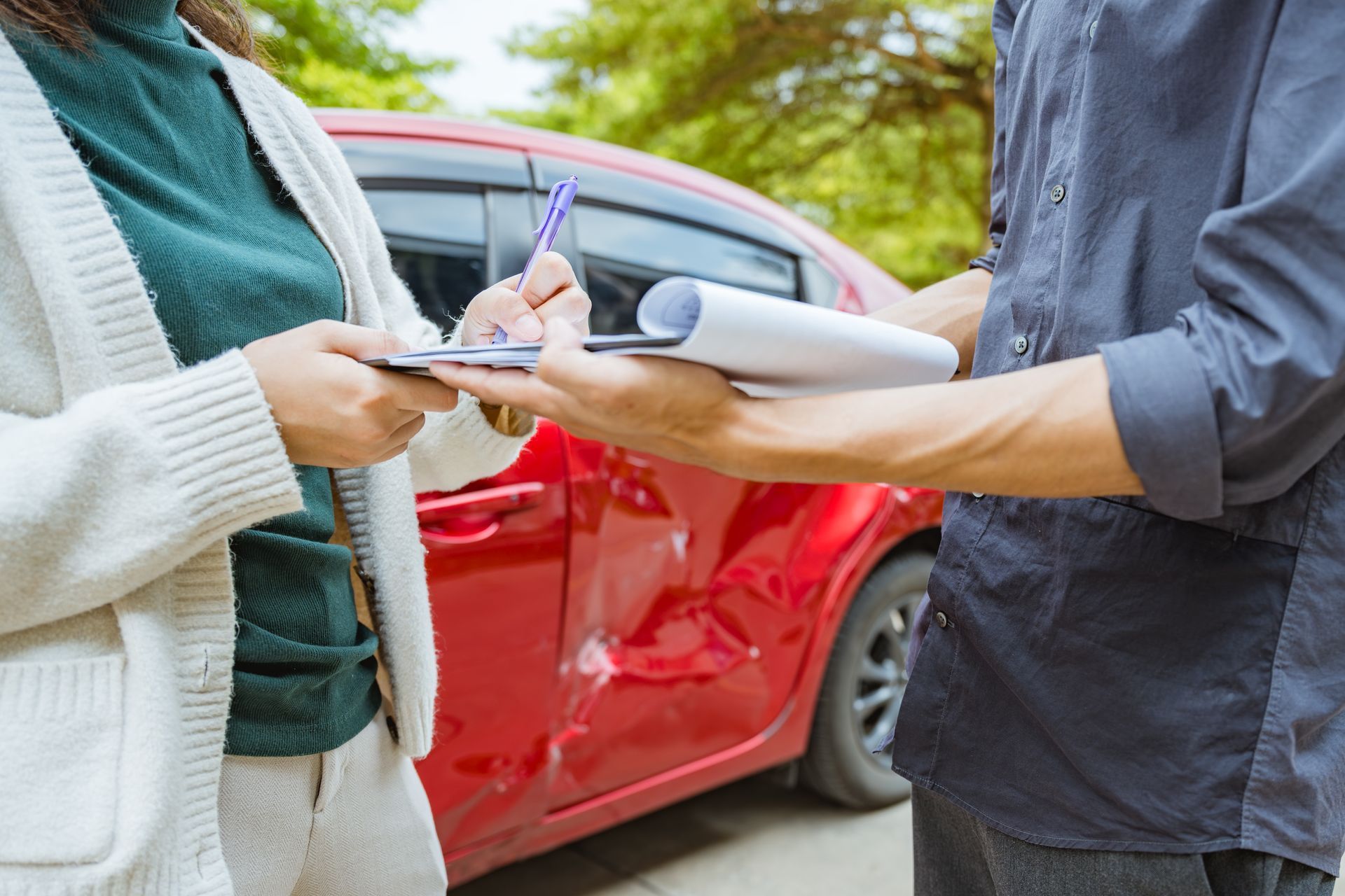 Two people exchanging car damage paperwork beside a red car after an accident