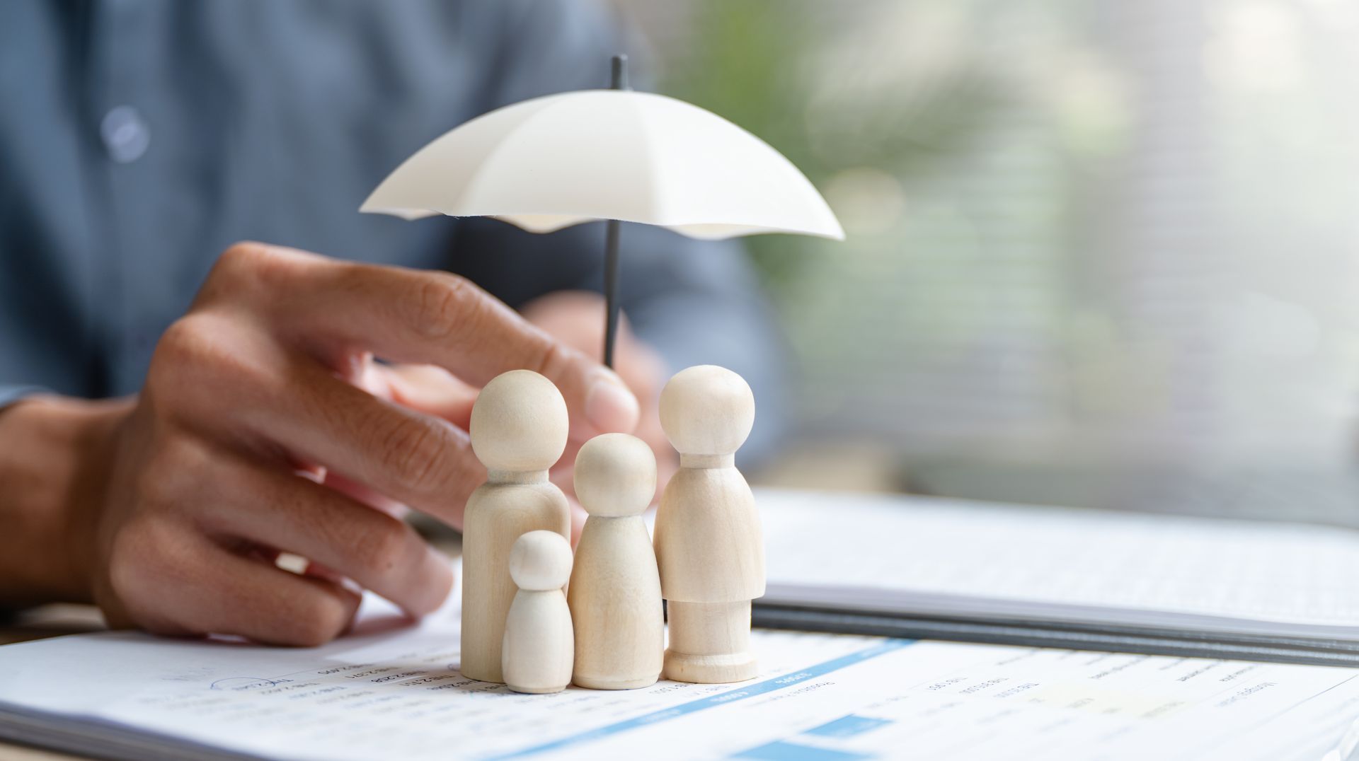 Hands arranging wooden figures under a white umbrella on a desk, symbolizing family insurance protection