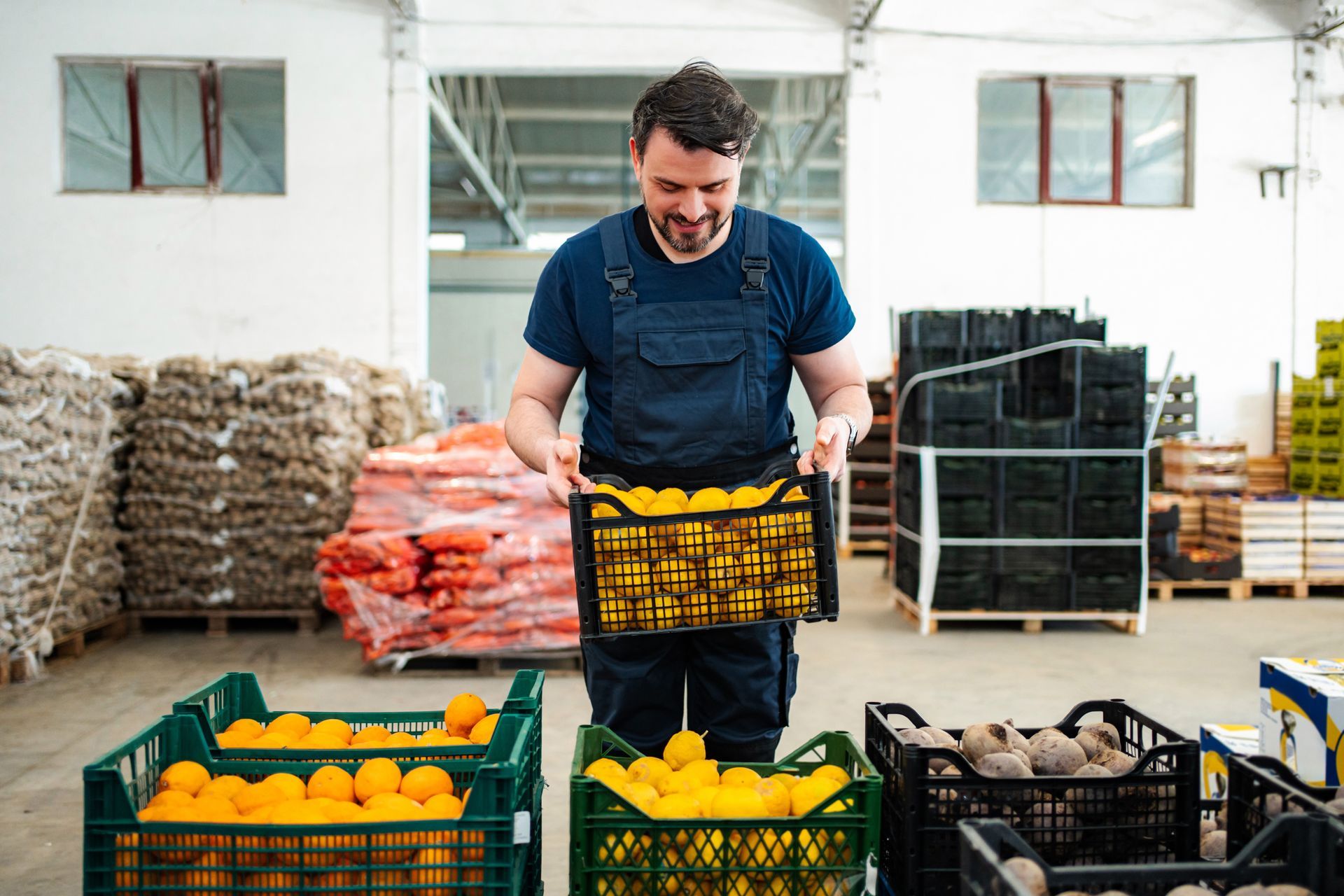 Man sorting produce crates in a warehouse, with oranges, vegetables, and stacked supplies around him.