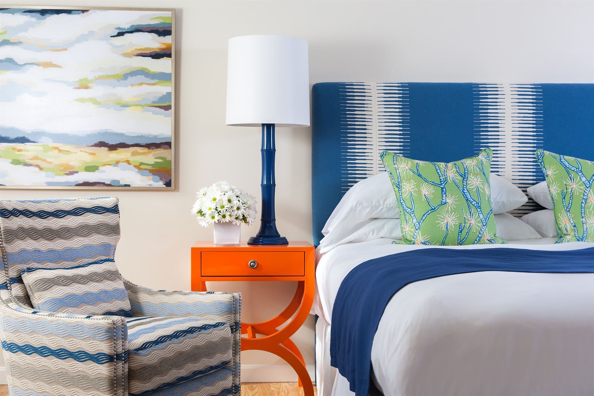 A bedroom with a bed , chair , nightstand , lamp and painting on the wall.