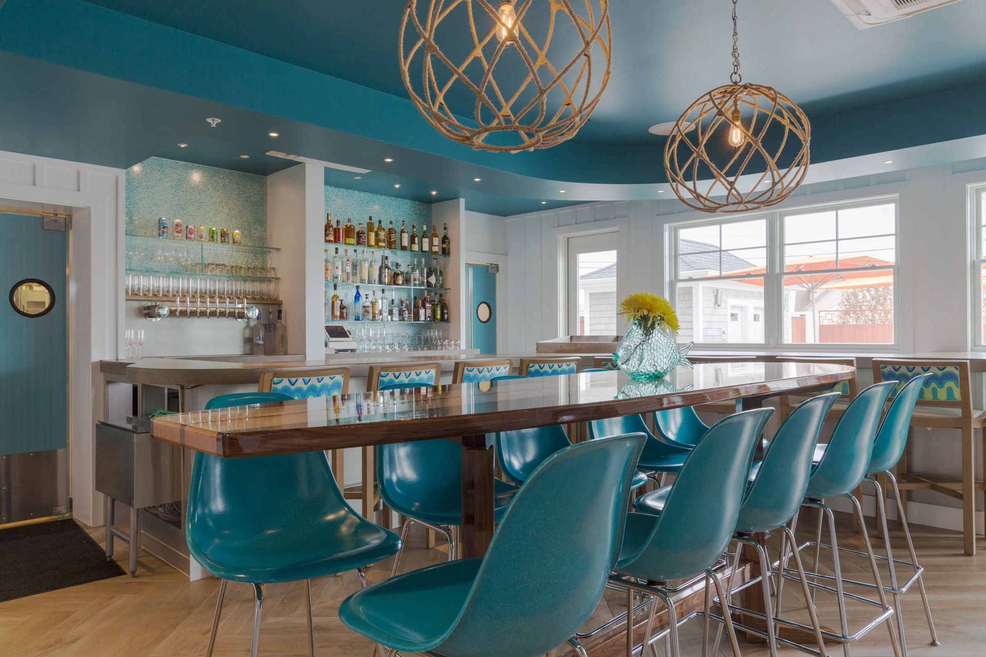 A restaurant with a long table and blue chairs and a bar.