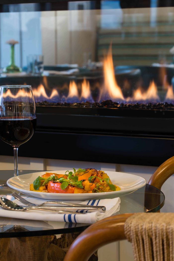 A plate of food and a glass of wine are on a table in front of a fireplace.