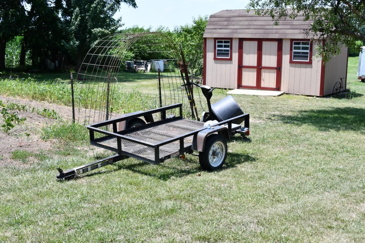 A trailer is parked in the grass in front of a shed.