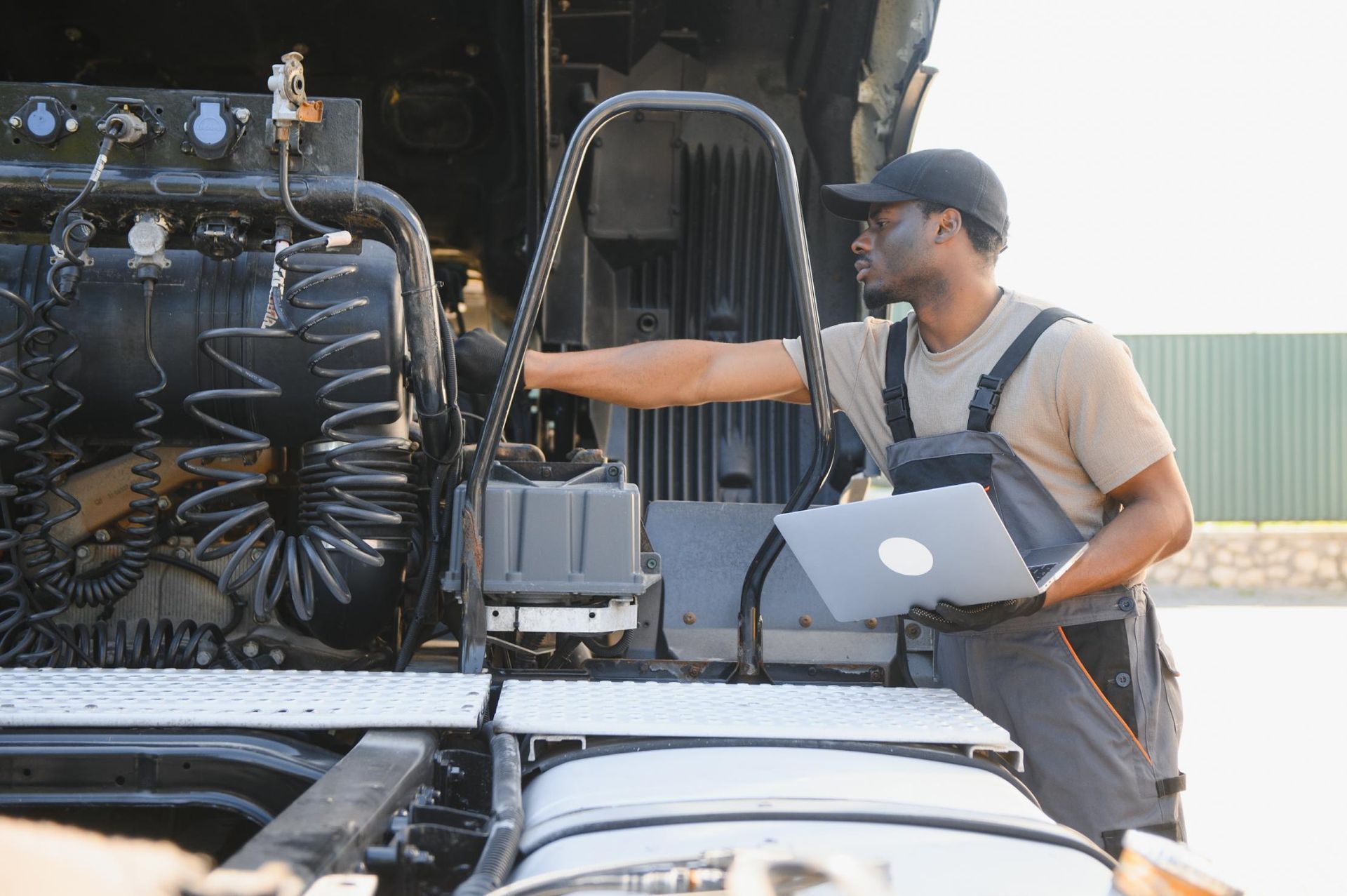 A man is working on the engine of a semi truck while holding a laptop.