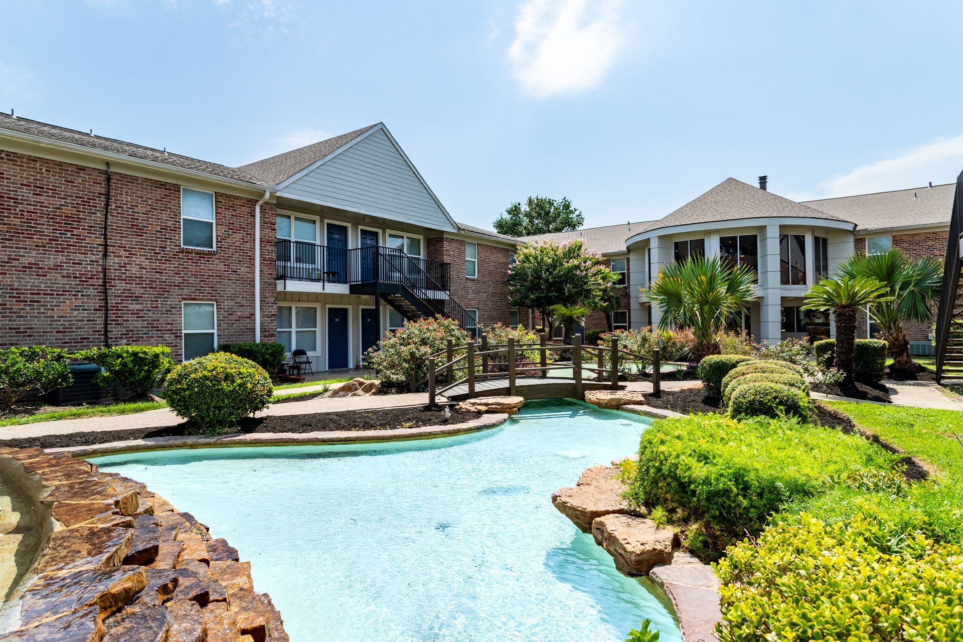 Apartment complex with a blue water feature, bridge, and lush landscaping under a blue sky.