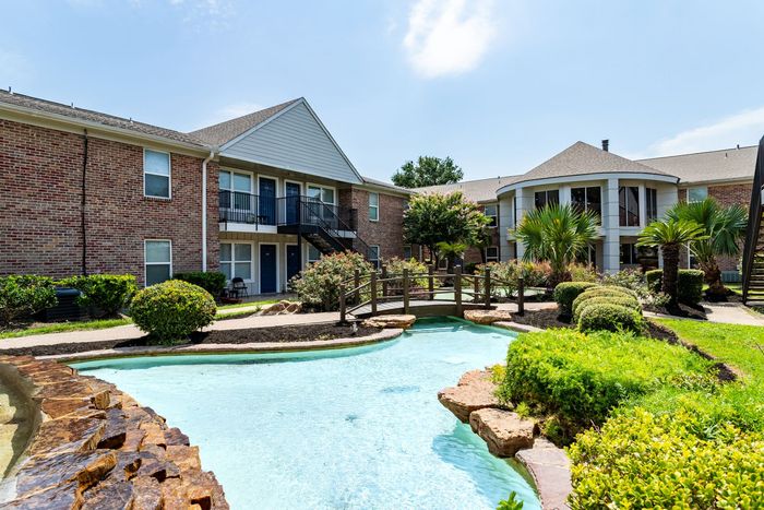 Apartment complex with a blue water feature, bridge, and lush landscaping under a blue sky.