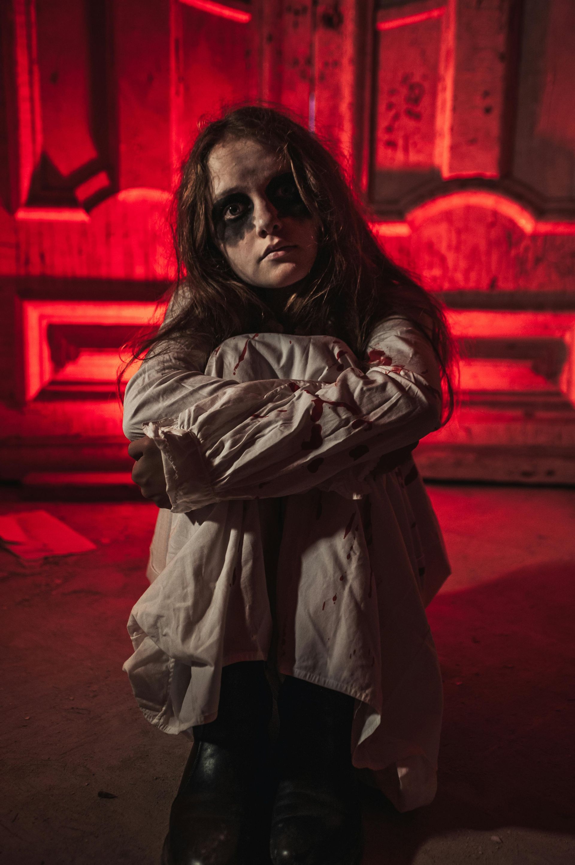 Girl in a white shirt, with dark eye makeup, sits in front of red doors, arms crossed, somber expression.