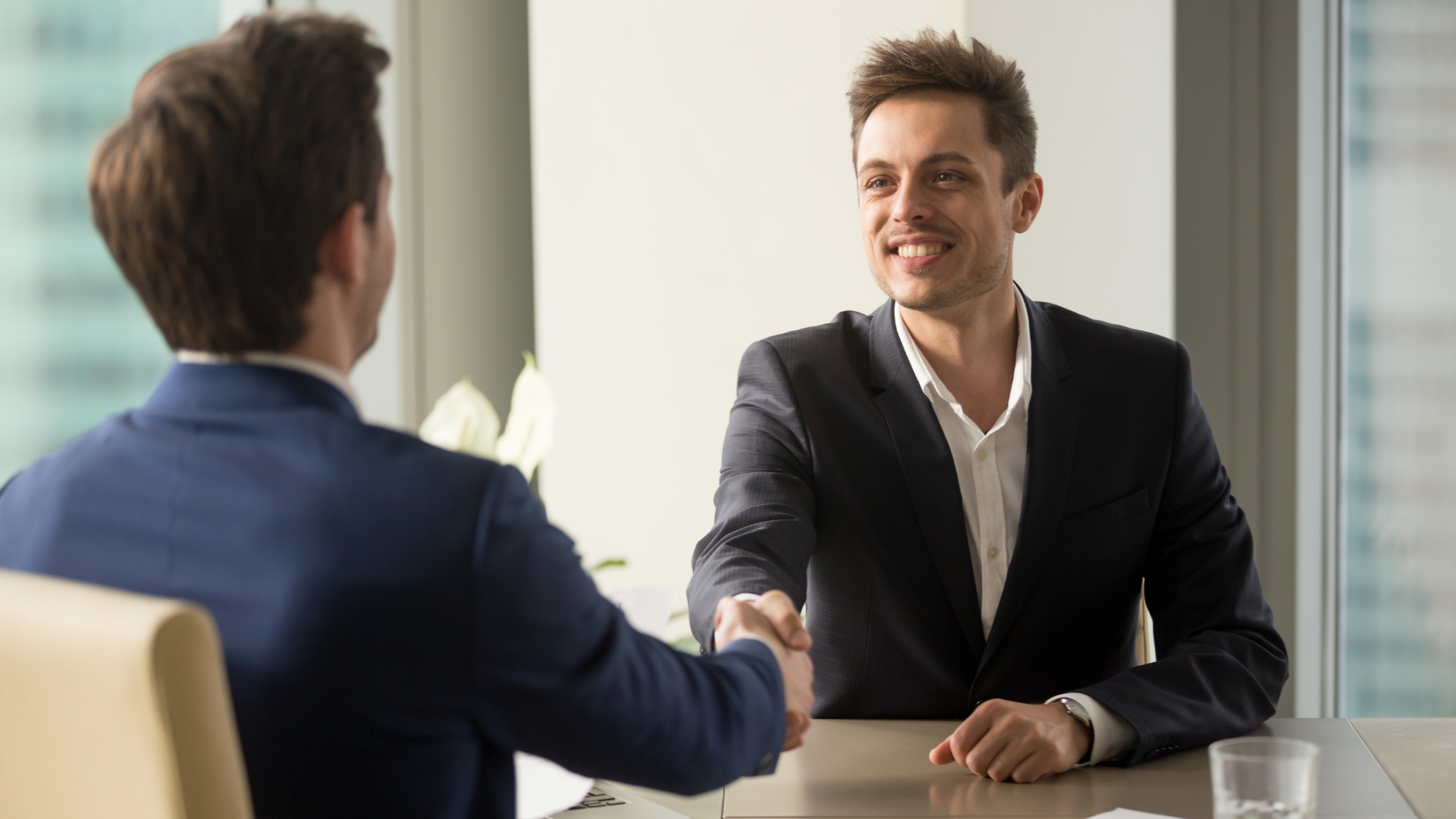 Two men in suits shaking hands across a table, smiling. Bright office setting.