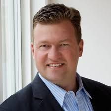 A man in a suit and striped shirt is smiling in front of a window.