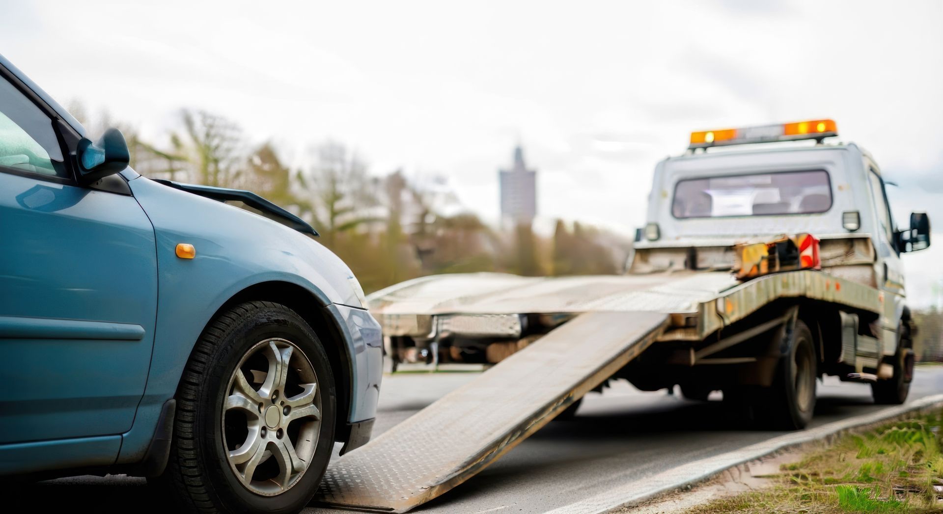 Damaged blue car in front of tow truck with ramp lowered on roadside.
