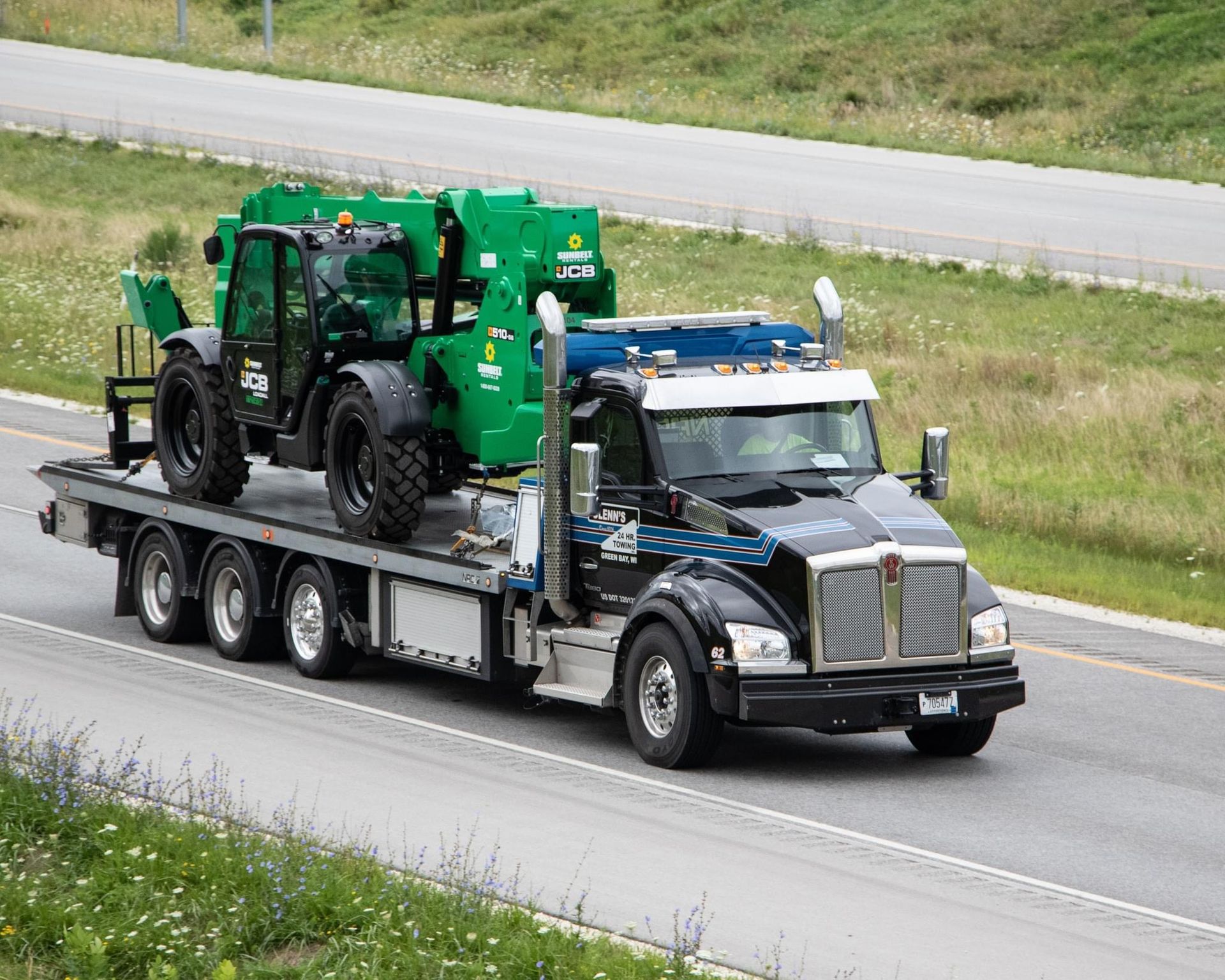 A black truck hauling a green forklift on a flatbed trailer, driving on a highway.