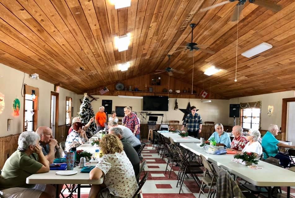 a group of people sit at long tables in a room with a ceiling fan