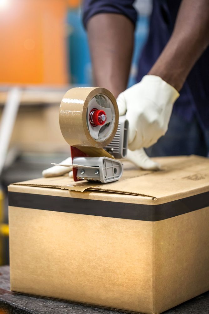 A Man is Taping a Cardboard Box With Tape — Wide Bay Removals In Dundowran, QLD A man is taping a cardboard box with tape — Wide Bay Removals In Dundowran, QLD