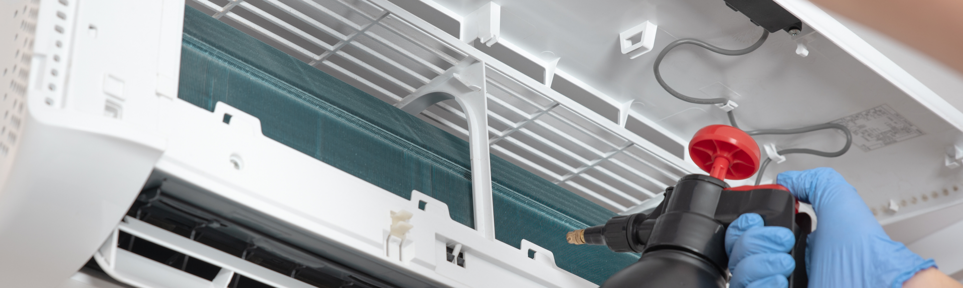 A person wearing blue gloves uses a pressurized spray bottle to clean the internal components of a wall-mounted air unit.