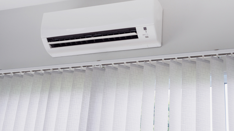 A wall-mounted white air conditioning unit installed above white vertical blinds.