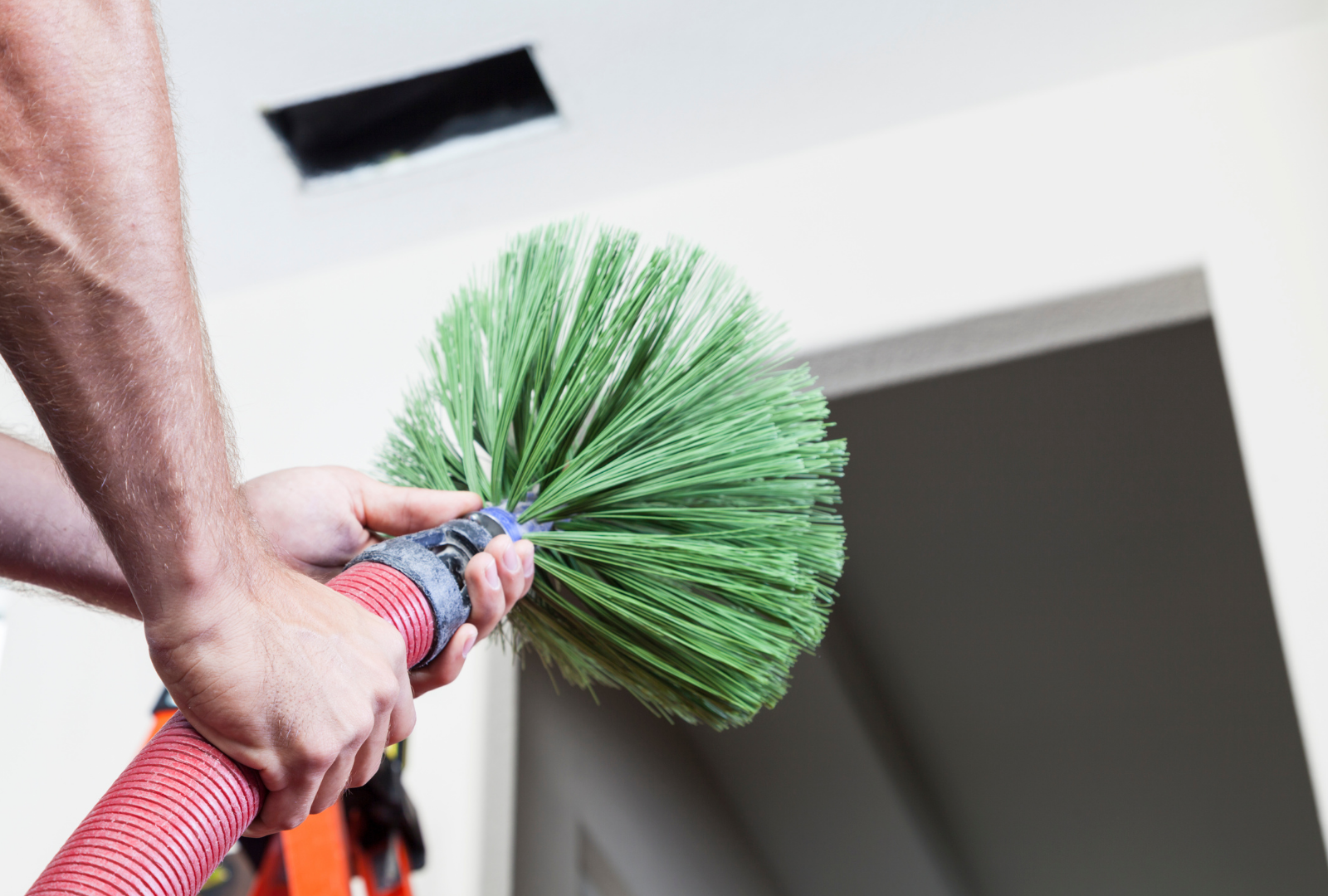 A person using a rotating green brush tool on an extension pole to clean a vent in a white ceiling.