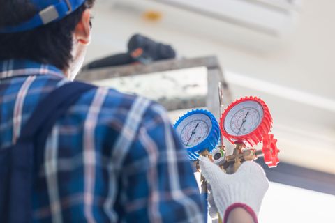 A technician wearing a blue plaid shirt and white gloves holds a set of HVAC pressure gauges while working on an AC unit.